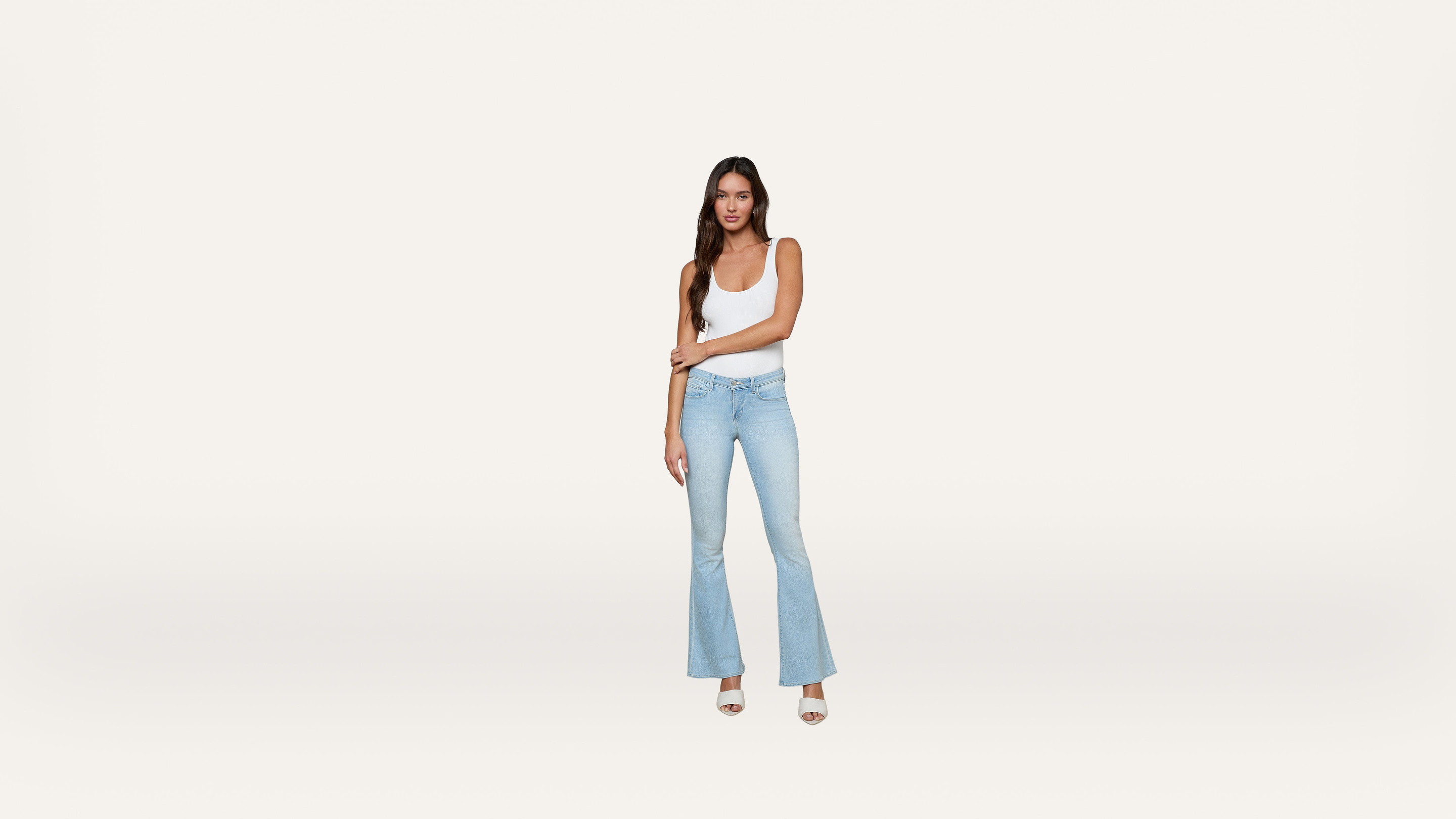 A woman in a white tank top and light blue flared jeans stands against a plain white background, looking at the camera with one arm crossed over her body.