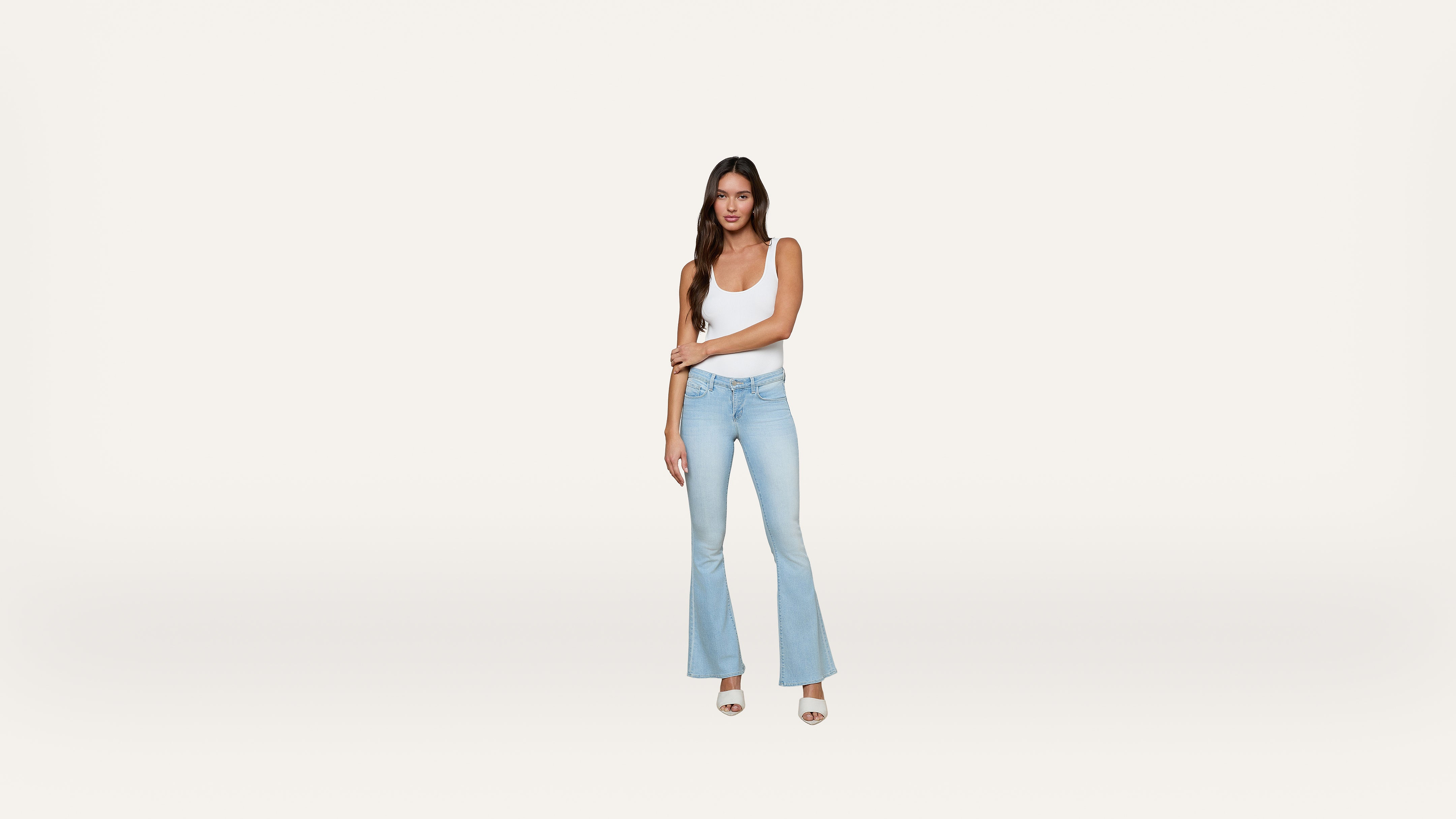 A woman in a white tank top and light blue flared jeans stands against a plain white background, looking at the camera with one arm crossed over her body.