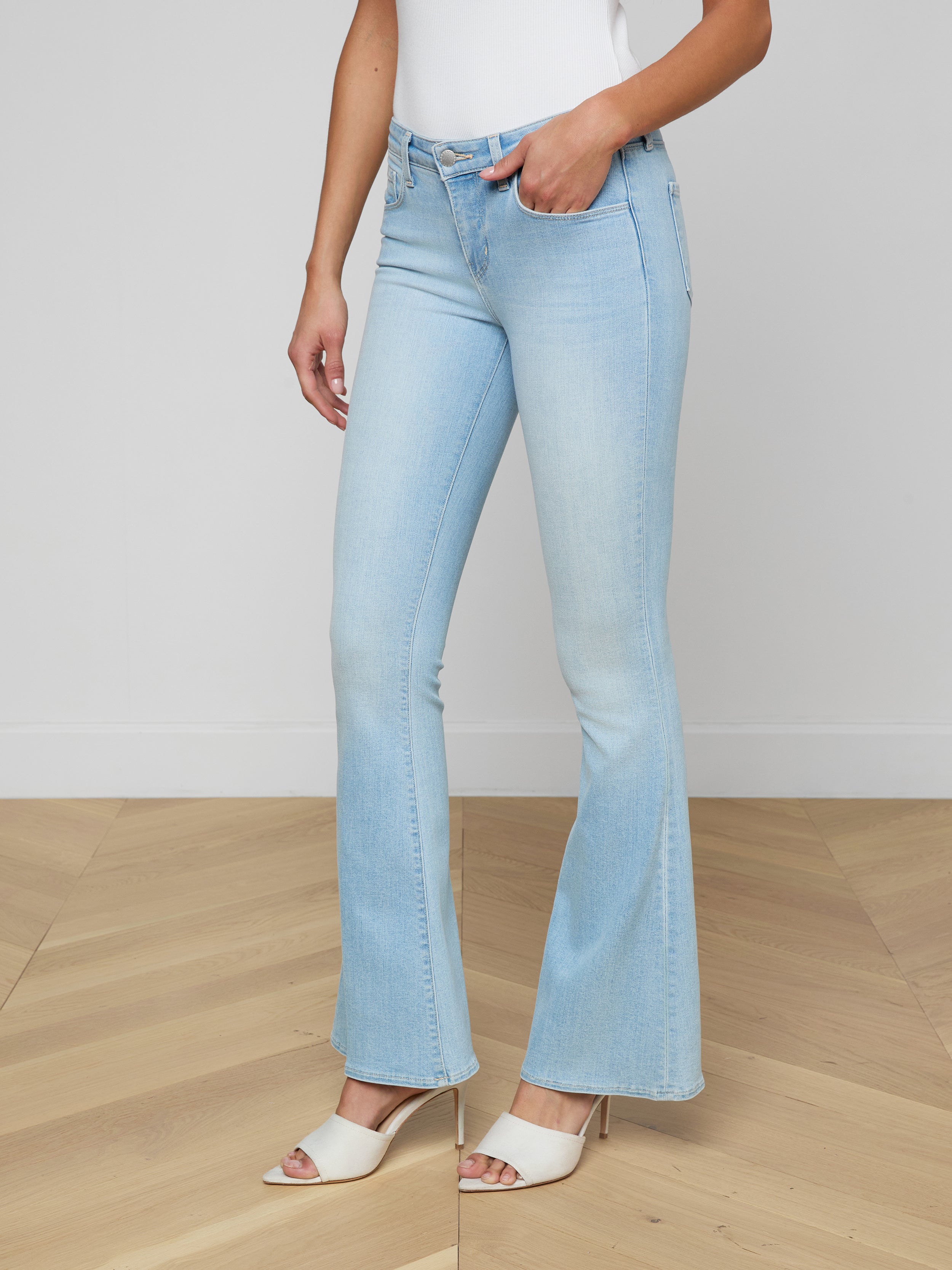 Wearing the Taney Low-Rise Flare Jean in light blue stretch denim, a person stands with one hand in their pocket, paired with a white sleeveless top and beige heeled sandals against a plain background.