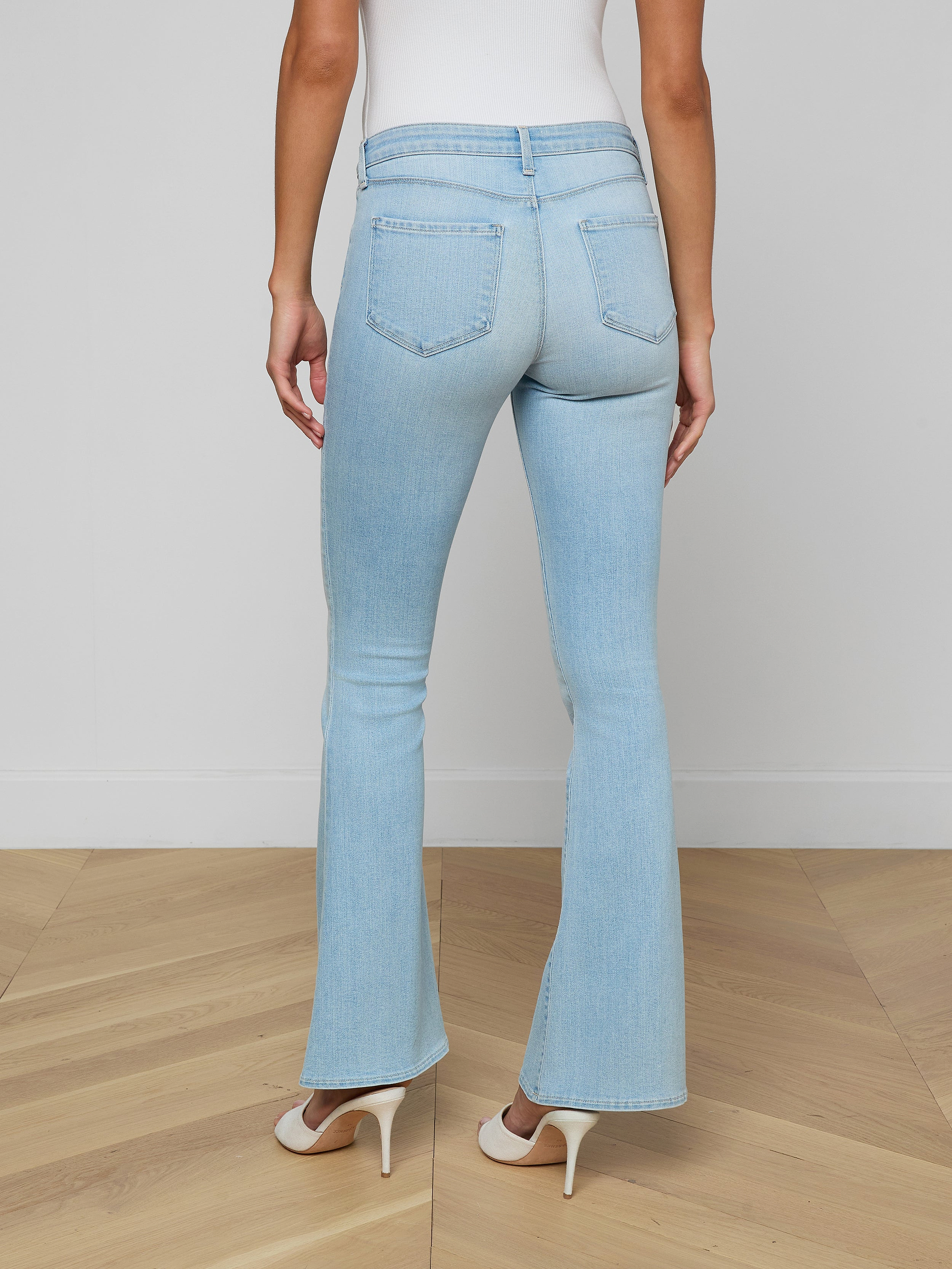 A woman stands facing away against a plain light background, showcasing the back of the Taney Low-Rise Flare Jean in light blue stretch denim, paired with white heels.