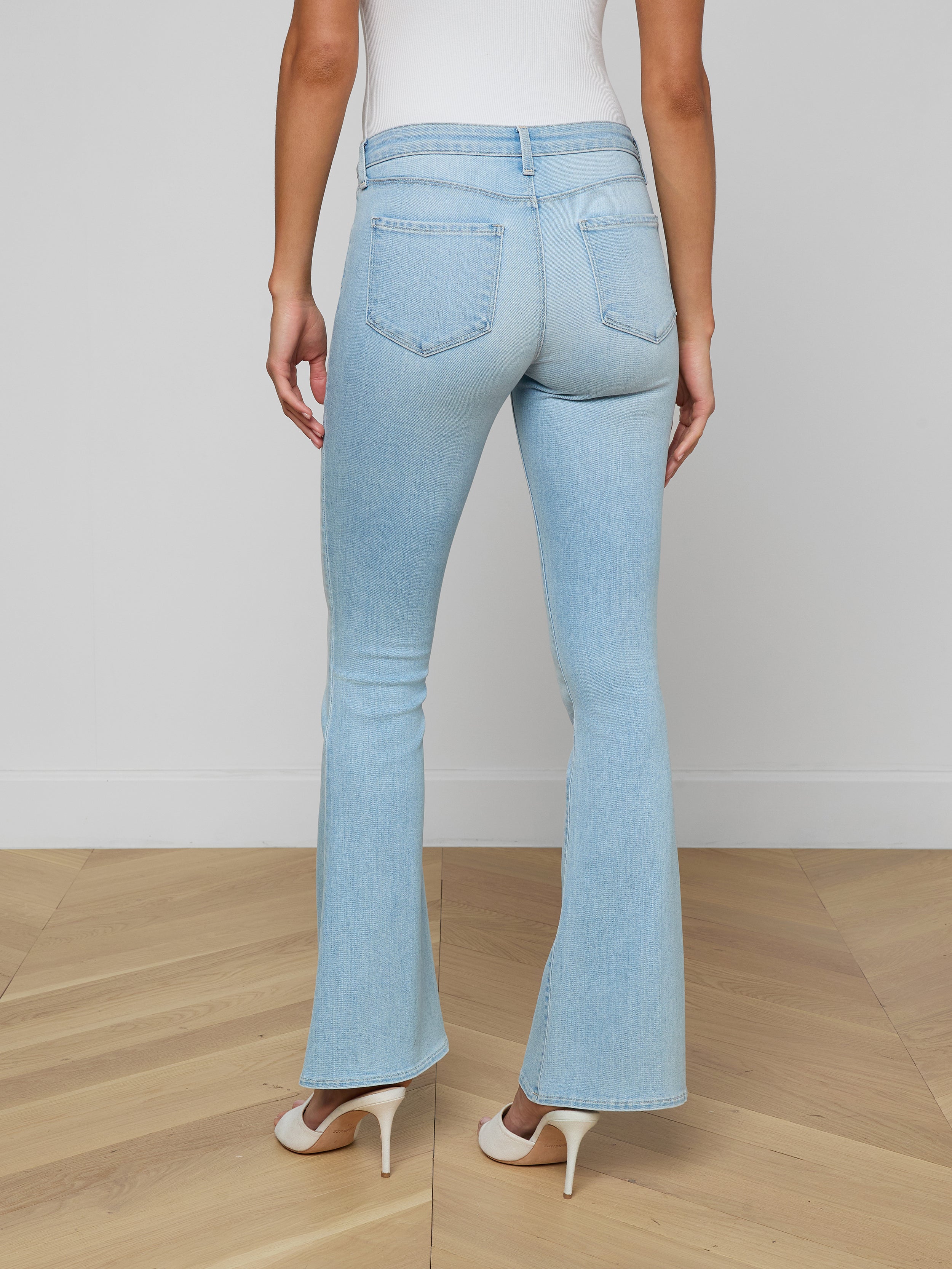 A woman stands facing away against a plain light background, showcasing the back of the Taney Low-Rise Flare Jean in light blue stretch denim, paired with white heels.