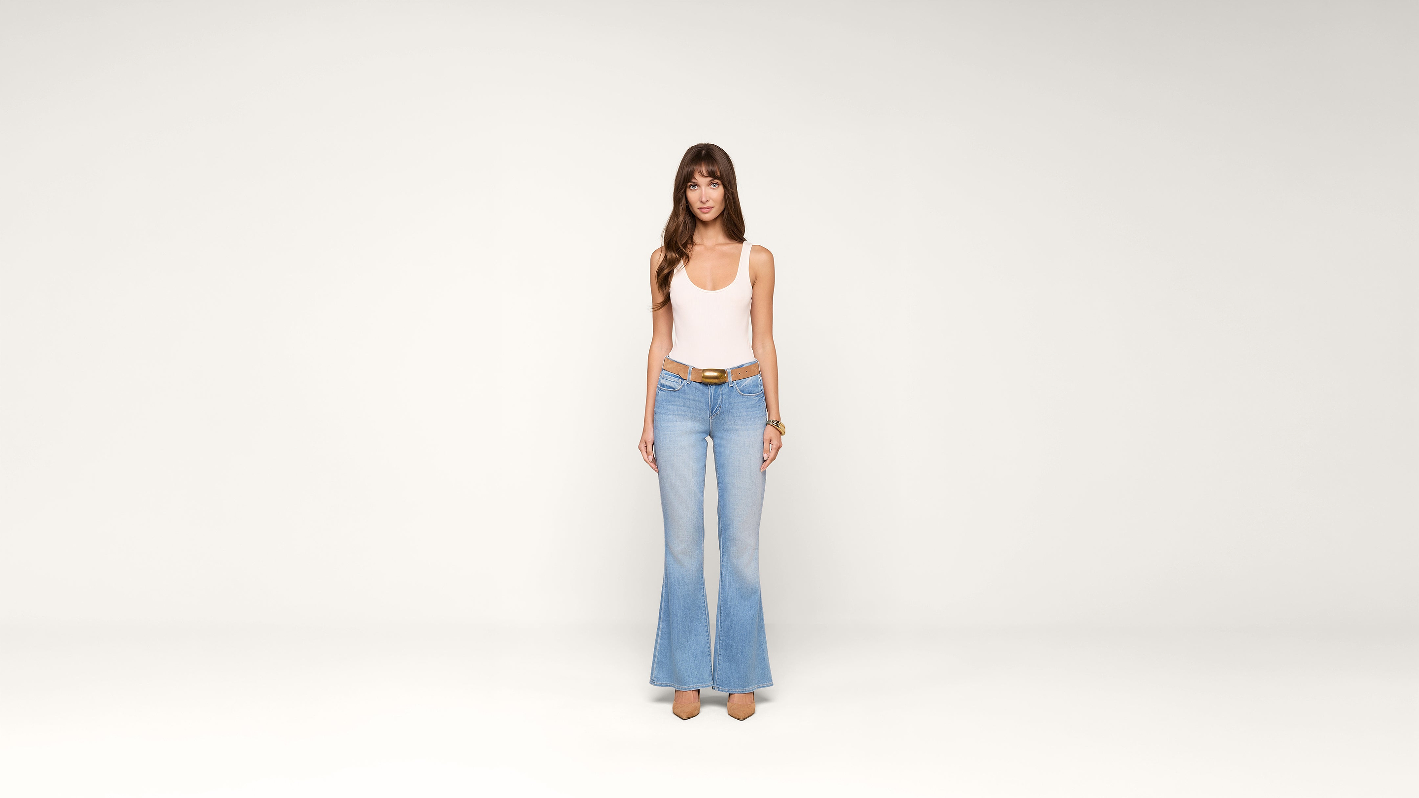 A woman wearing a white tank top, light blue flared jeans, and beige shoes stands facing forward against a plain white background.