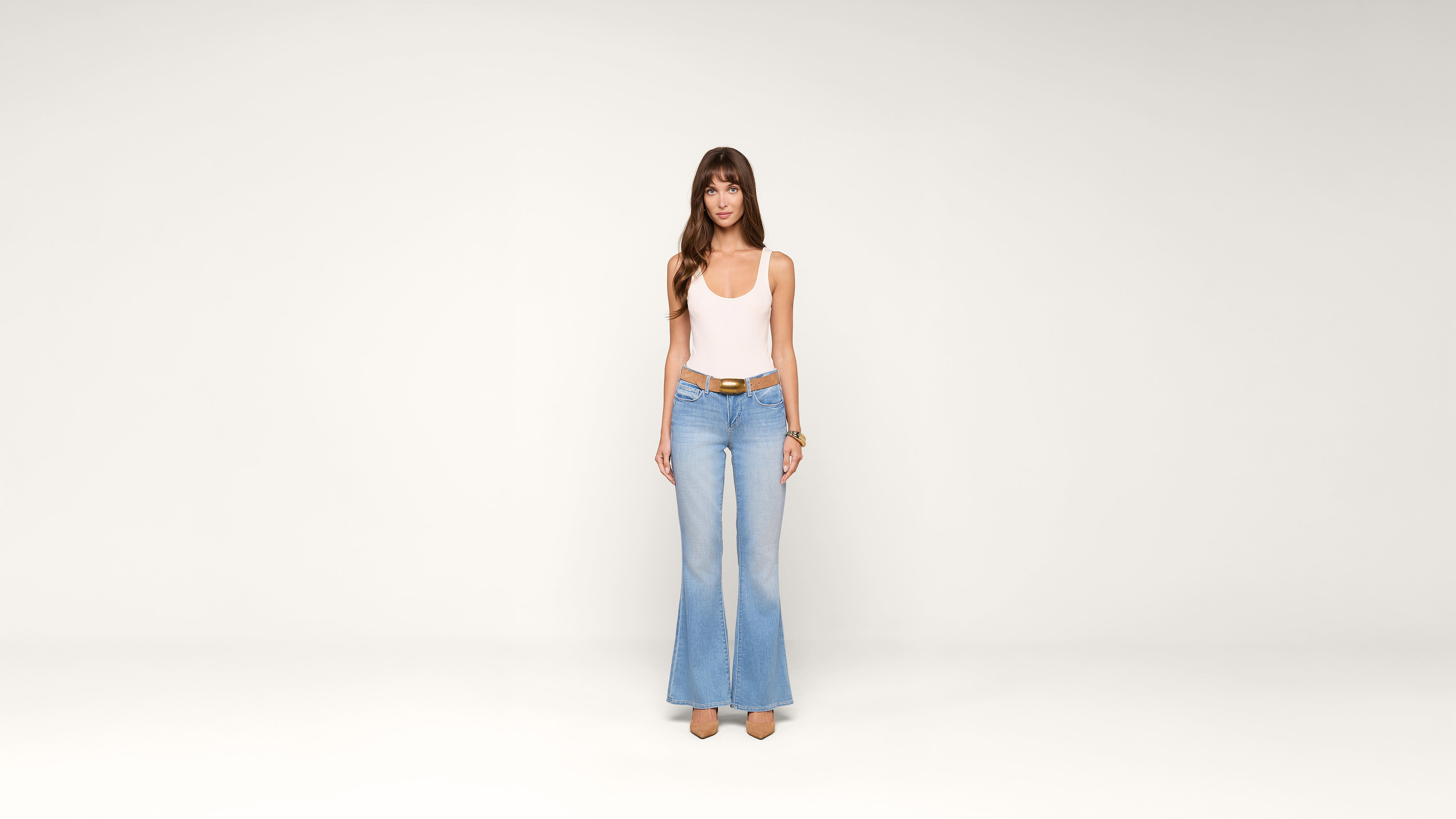 A woman wearing a white tank top, light blue flared jeans, and beige shoes stands facing forward against a plain white background.