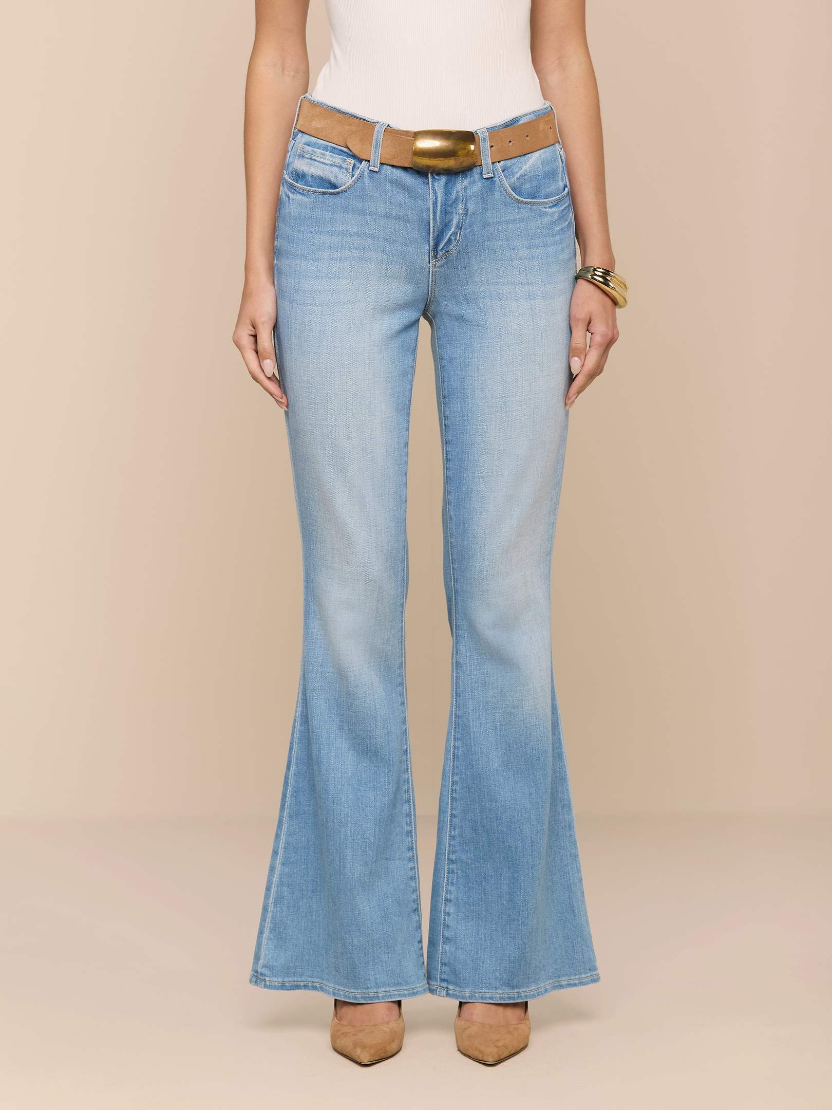 Vêtue dun jean évasé à taille basse Taney en délavage indigo clair, dune ceinture beige à boucle en laiton, dun haut blanc, de chaussures beiges à bout pointu et dun bracelet doré, cette personne se détache sur un fond neutre.