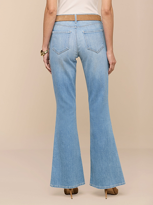 A person is seen from the back wearing the Taney Low-Rise Flare Jean in light indigo wash, styled with tan high heels, a white top, and bracelets, against a beige background. The jeans feature two back pockets.