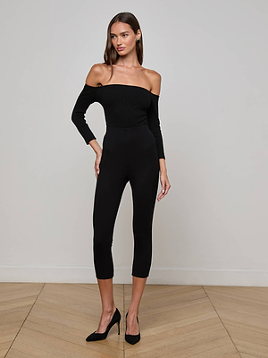 A woman stands indoors against a light gray wall, wearing the Dee Capri Pant with black heels. She has long brown hair and poses with one hand on her hip on a wooden floor.