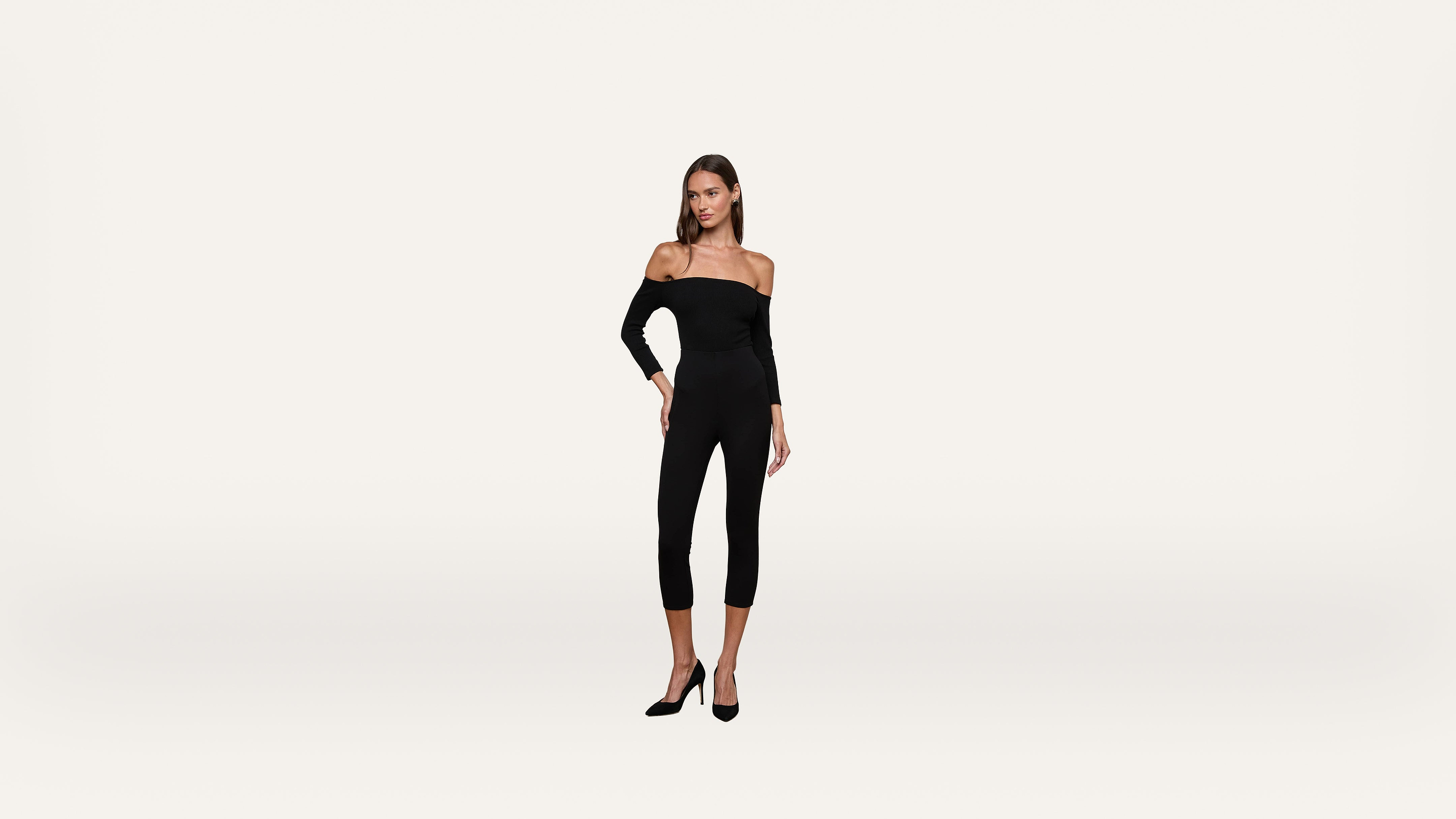 A woman wearing a black off-the-shoulder jumpsuit and black heels stands against a plain white background.