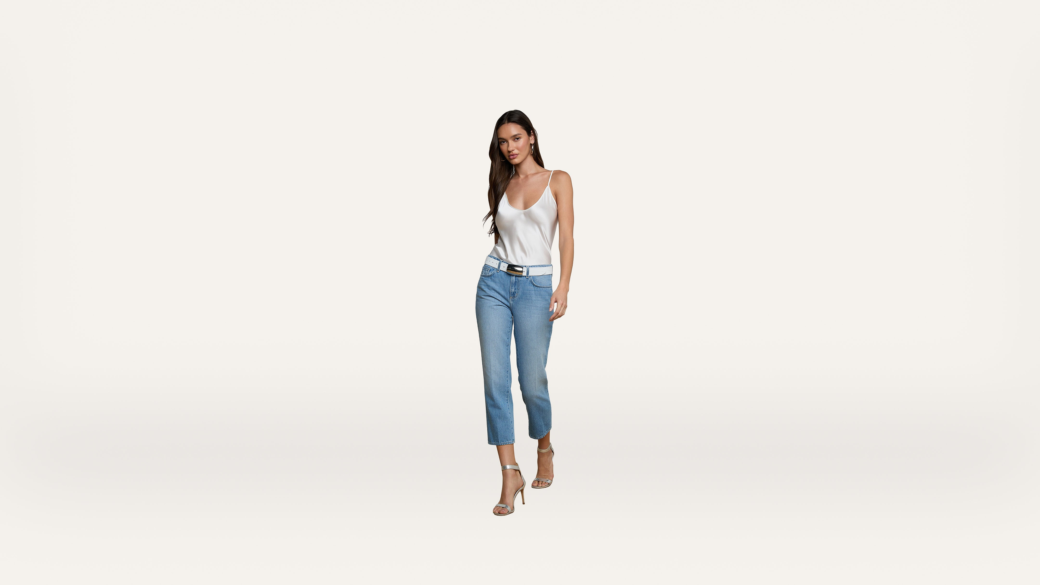 A woman with long dark hair is wearing a white tank top, blue jeans, and heels, walking confidently against a plain white background.