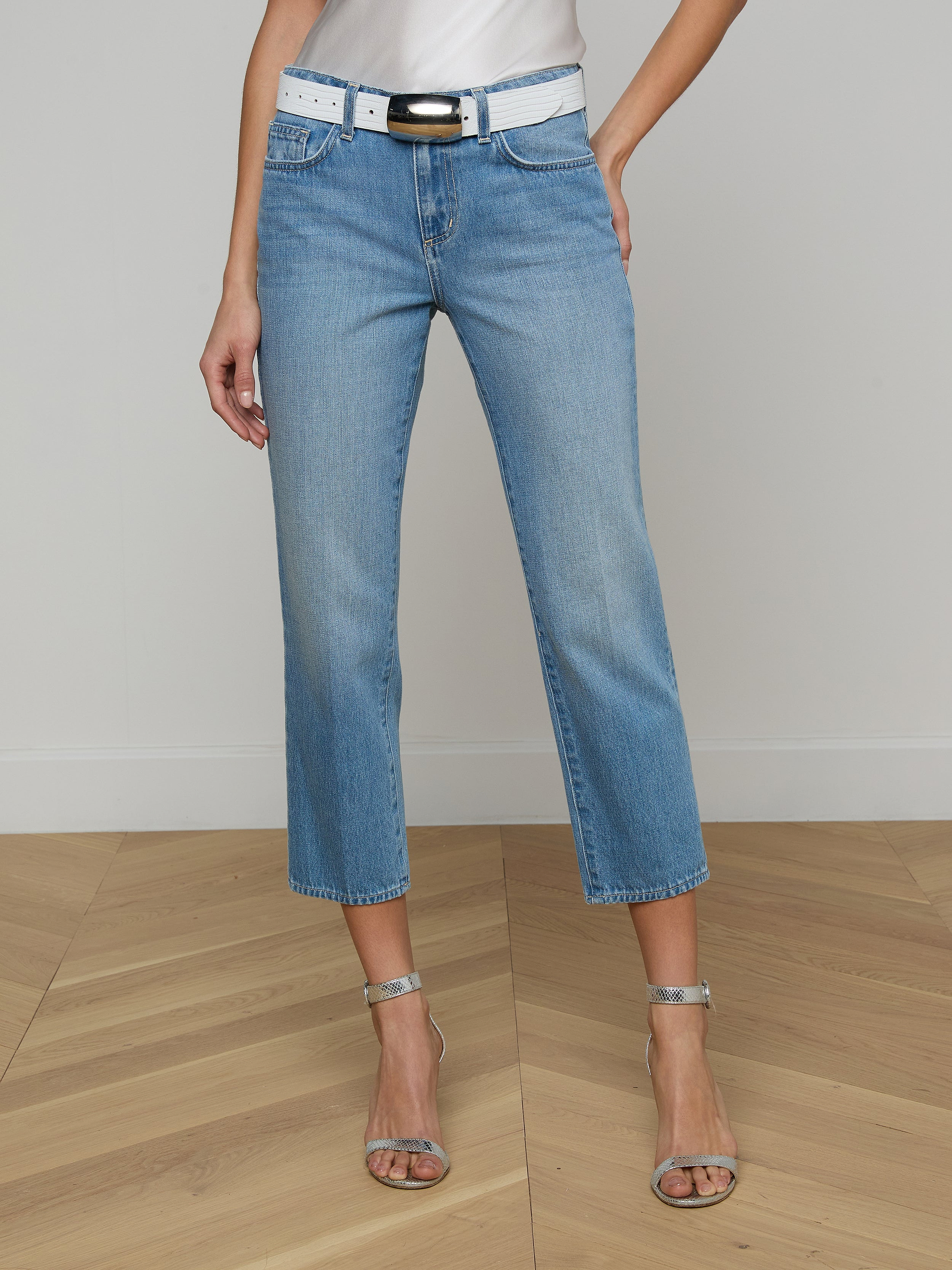 A person wearing the Thalina Cropped Stovepipe Jean in a light blue wash, paired with a white belt, a white top, and silver high-heeled sandals stands against a plain light background.