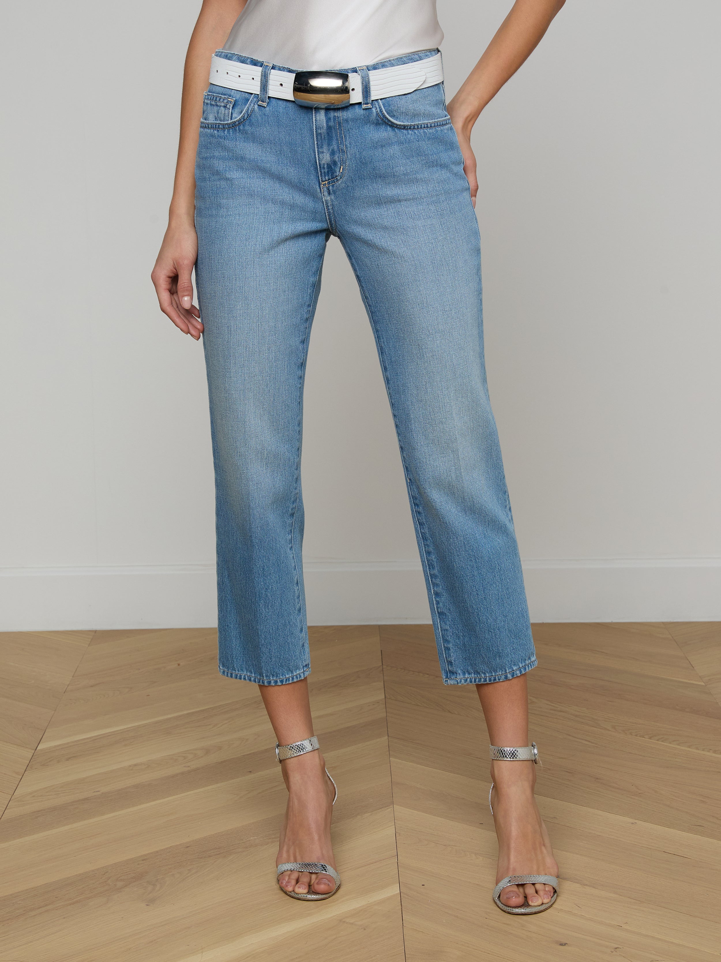A person wearing the Thalina Cropped Stovepipe Jean in a light blue wash, paired with a white belt, a white top, and silver high-heeled sandals stands against a plain light background.