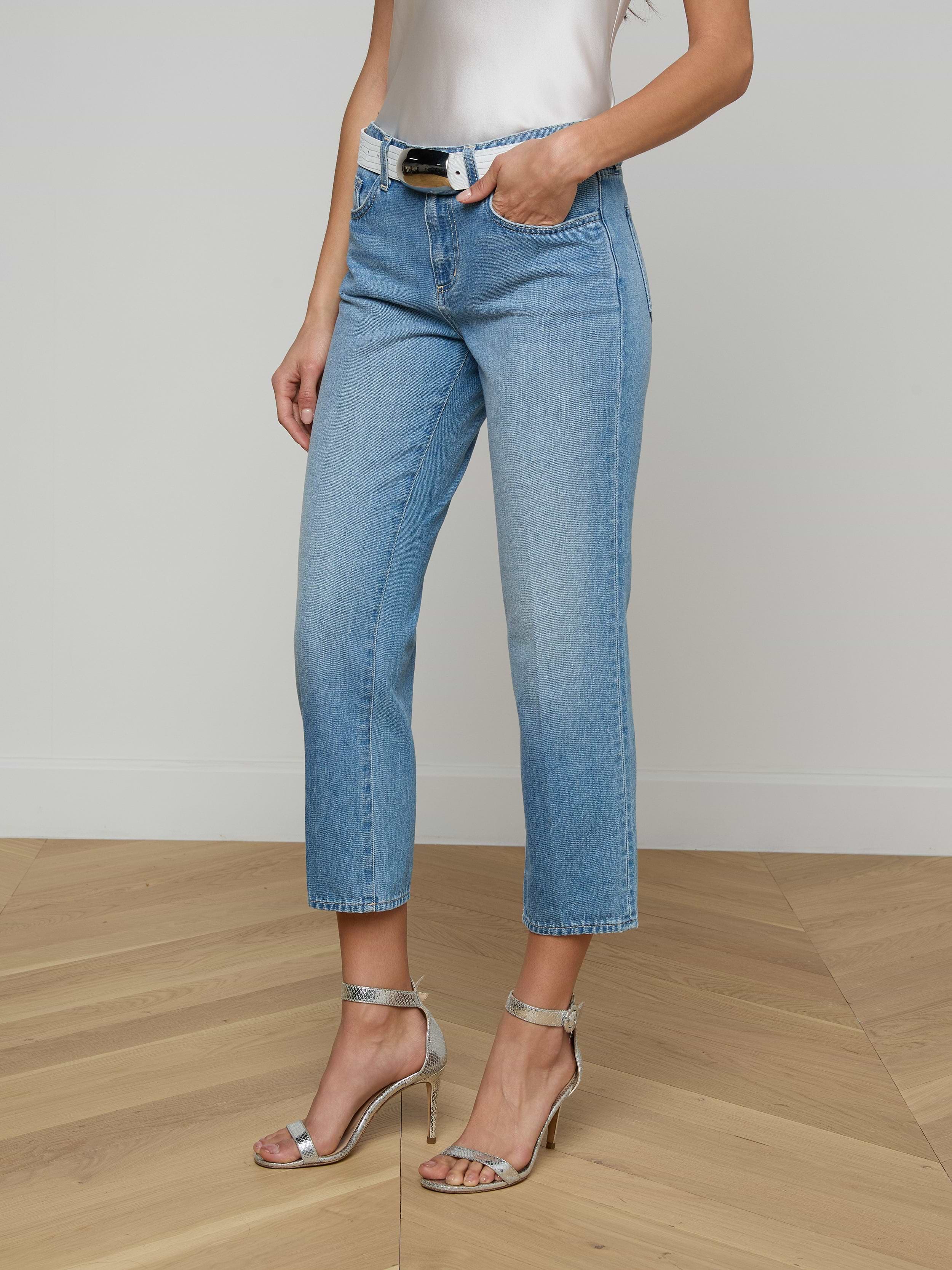 A woman stands with one hand in her pocket against a plain background, wearing the Thalina Cropped Stovepipe Jean in a light blue wash, paired with a white sleeveless top, black belt, and silver high-heeled sandals.