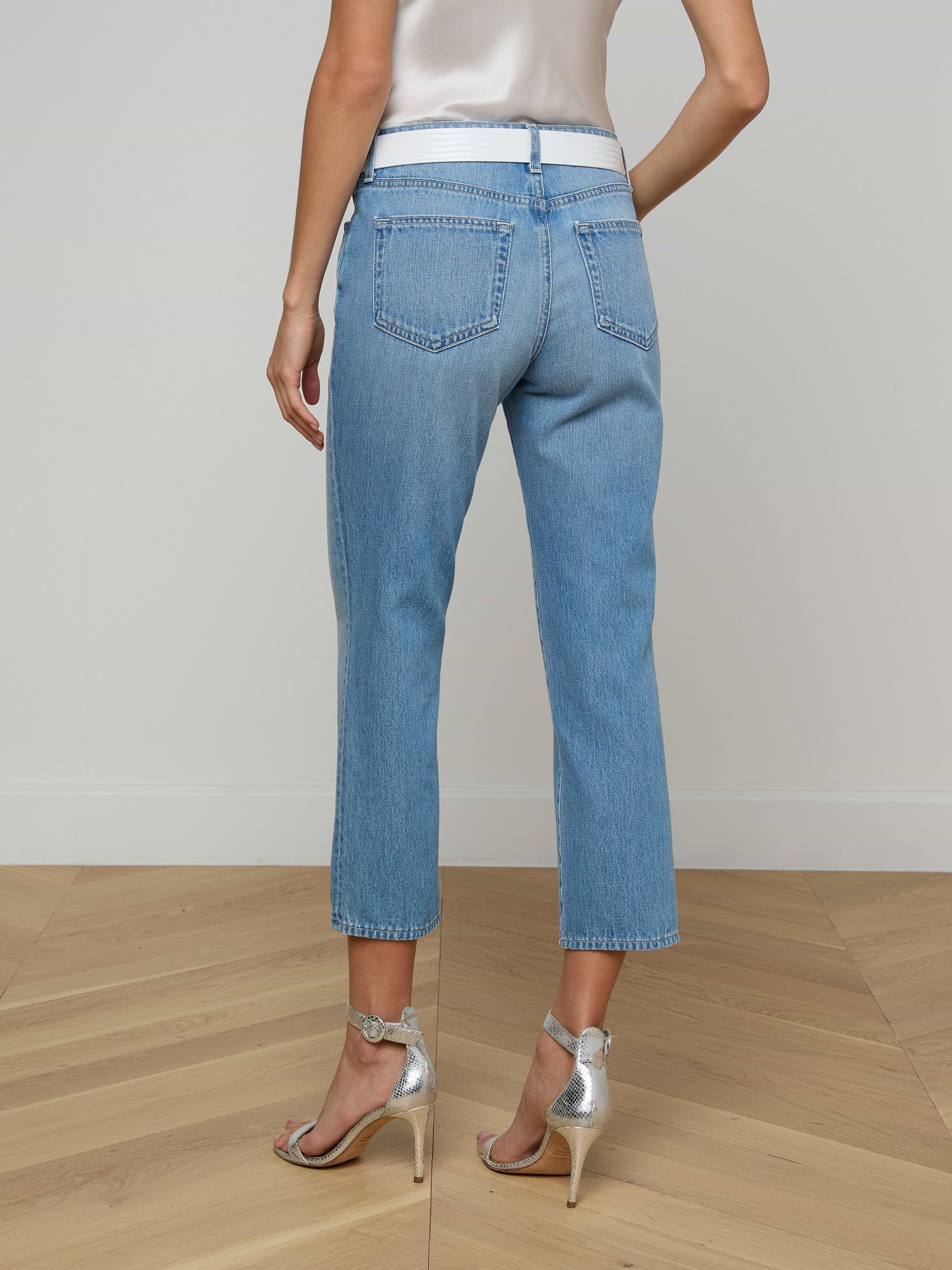 A woman stands with her back facing the camera against a plain white background, wearing the Thalina Cropped Stovepipe Jean in a light blue wash and silver high heels.