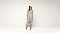 A woman stands in a light gray room wearing a matching light gray crop top and loose, high-waisted pants with ankle ties, paired with strappy heels.