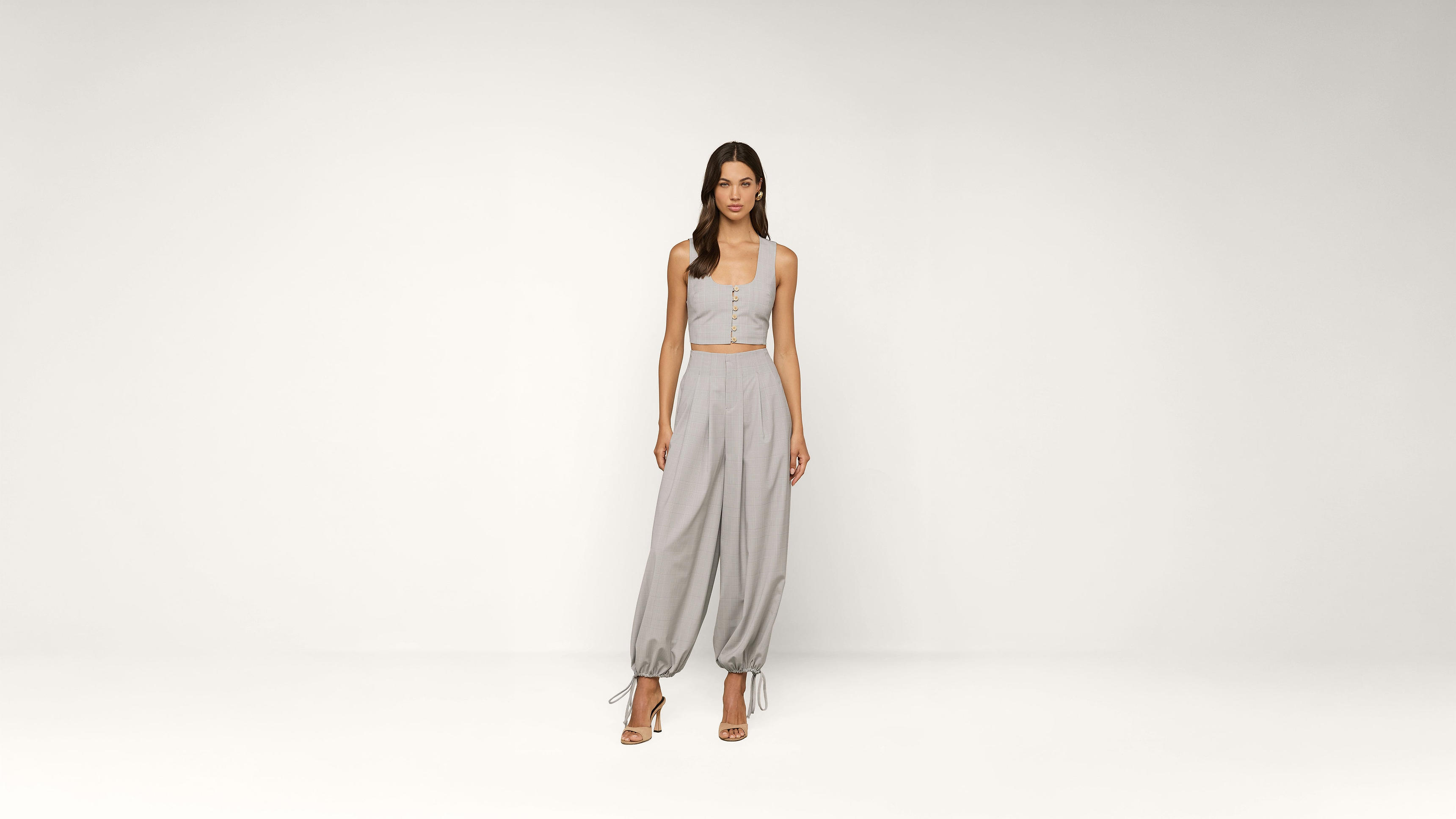 A woman stands in a light gray room wearing a matching light gray crop top and loose, high-waisted pants with ankle ties, paired with strappy heels.
