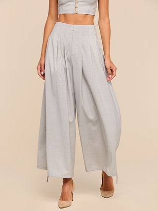 A woman wears the Ysola Pant, featuring an ultra-high rise and wide-leg silhouette in a light gray plaid luxurious wool blend with pleats. She pairs them with nude heels and a cropped top against a beige background.