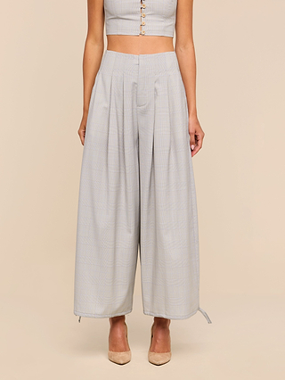 A woman wears the Ysola Pant, an ultra-high rise, wide-leg trouser in light gray with pleats, crafted from a soft wool blend. Styled with a cropped top and beige pointed-toe heels, her hands rest naturally at her sides against a neutral background.