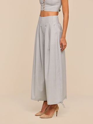 A woman stands against a plain beige background wearing the Ysola Pant—ultra-high rise, wide-leg, light gray trousers crafted from a soft wool blend—paired with a matching sleeveless buttoned crop top and beige pointed-toe heels.