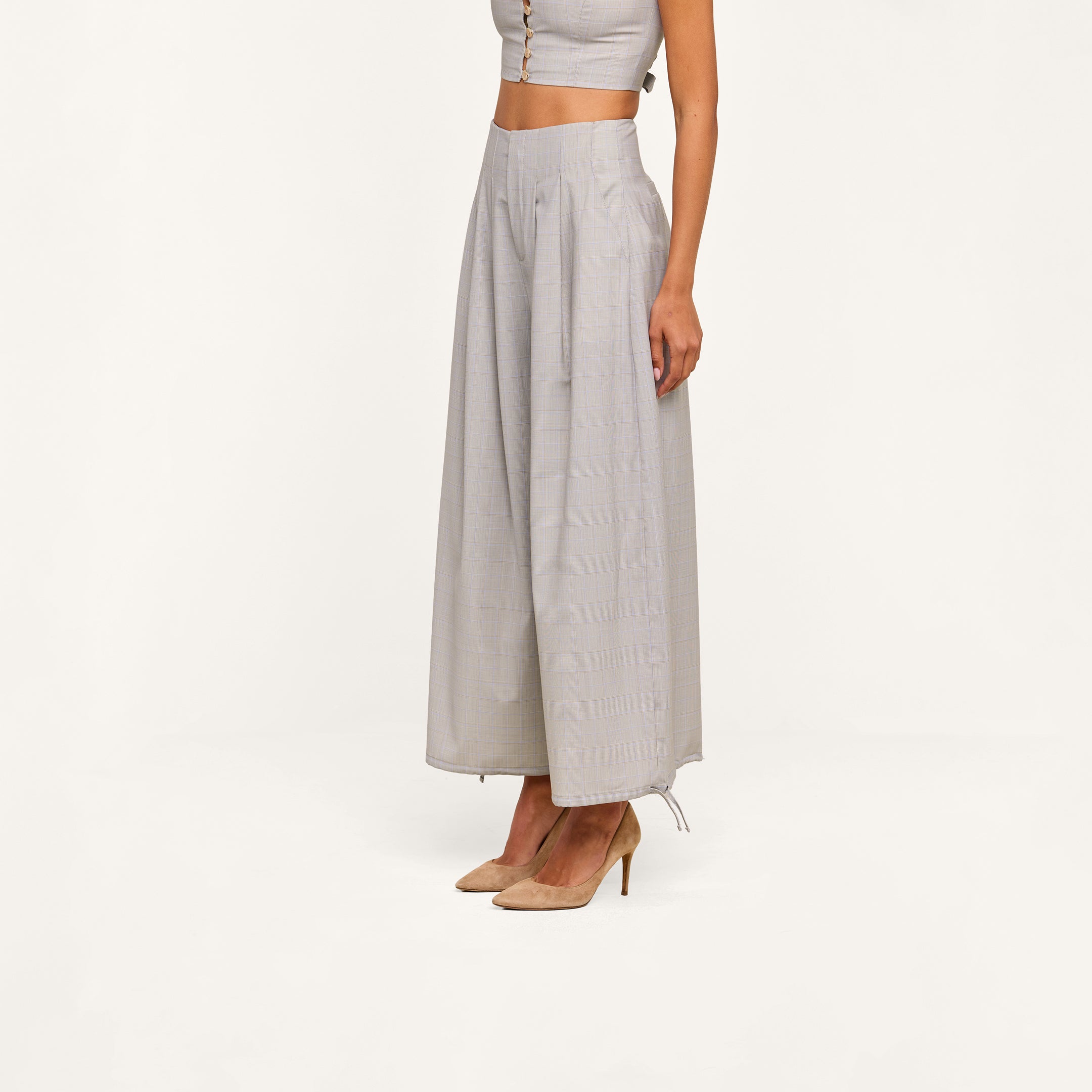 A woman wearing a sleeveless crop top and the Ysola Pant—ultra-high rise, wide-leg, wool blend light gray pants with pleats—paired with beige high-heeled shoes, stands against a plain white background.