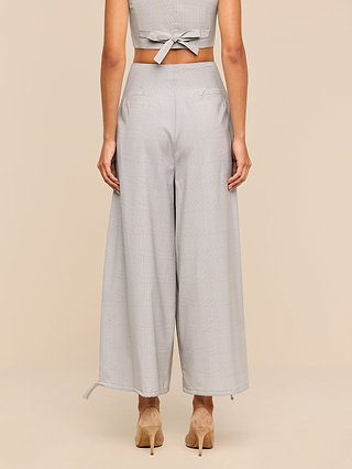 A woman is seen from behind, wearing the Ysola Pant in light gray wool blend with a wide-leg silhouette, paired with a matching sleeveless tie-back top and beige high-heeled shoes, set against a plain beige background.