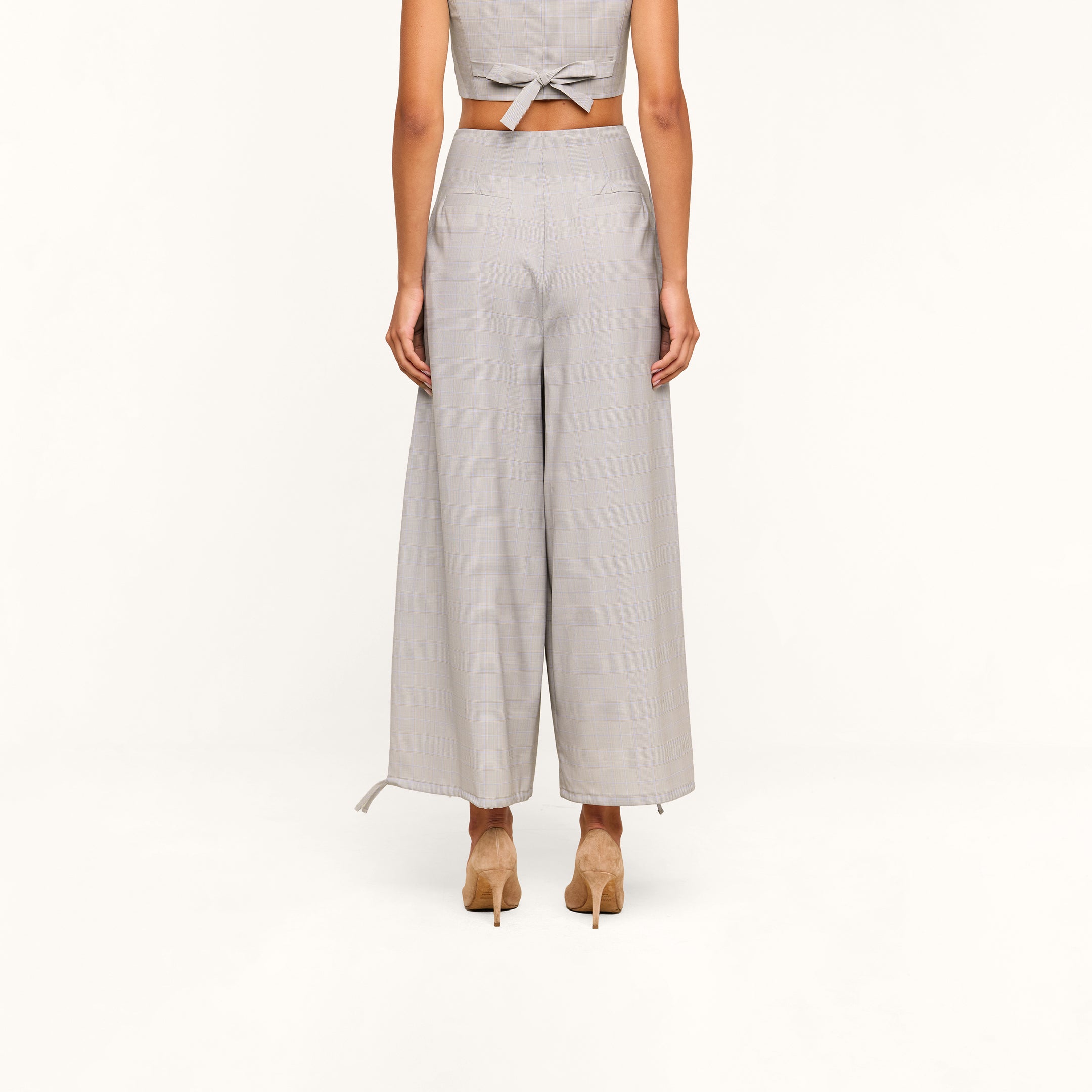 A person stands facing away, wearing the Ysola Pant—an ankle-length, ultra-high-rise style in a light gray wool blend with wide legs and a partially visible back tie detail at the top. Beige heels complete the look against a plain white background.