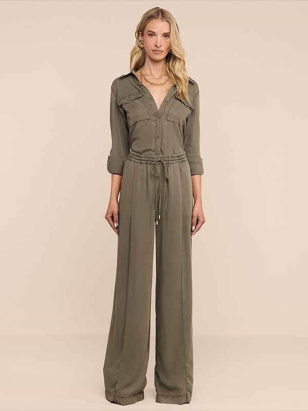 Una donna è rivolta in avanti e indossa i Connie Drawstring Trouser in verde oliva con coulisse in vita e pantaloni a gamba larga, su uno sfondo beige.