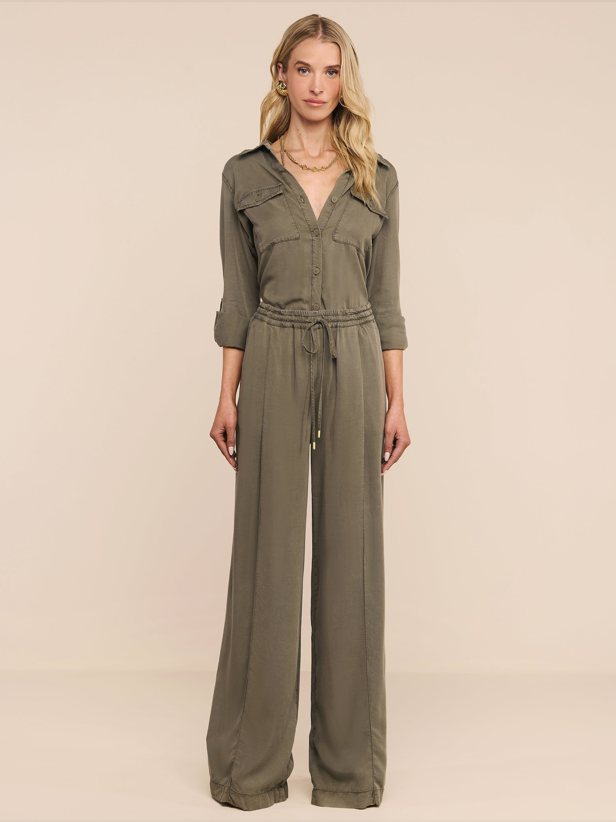 Una donna è rivolta in avanti e indossa i Connie Drawstring Trouser in verde oliva con coulisse in vita e pantaloni a gamba larga, su uno sfondo beige.