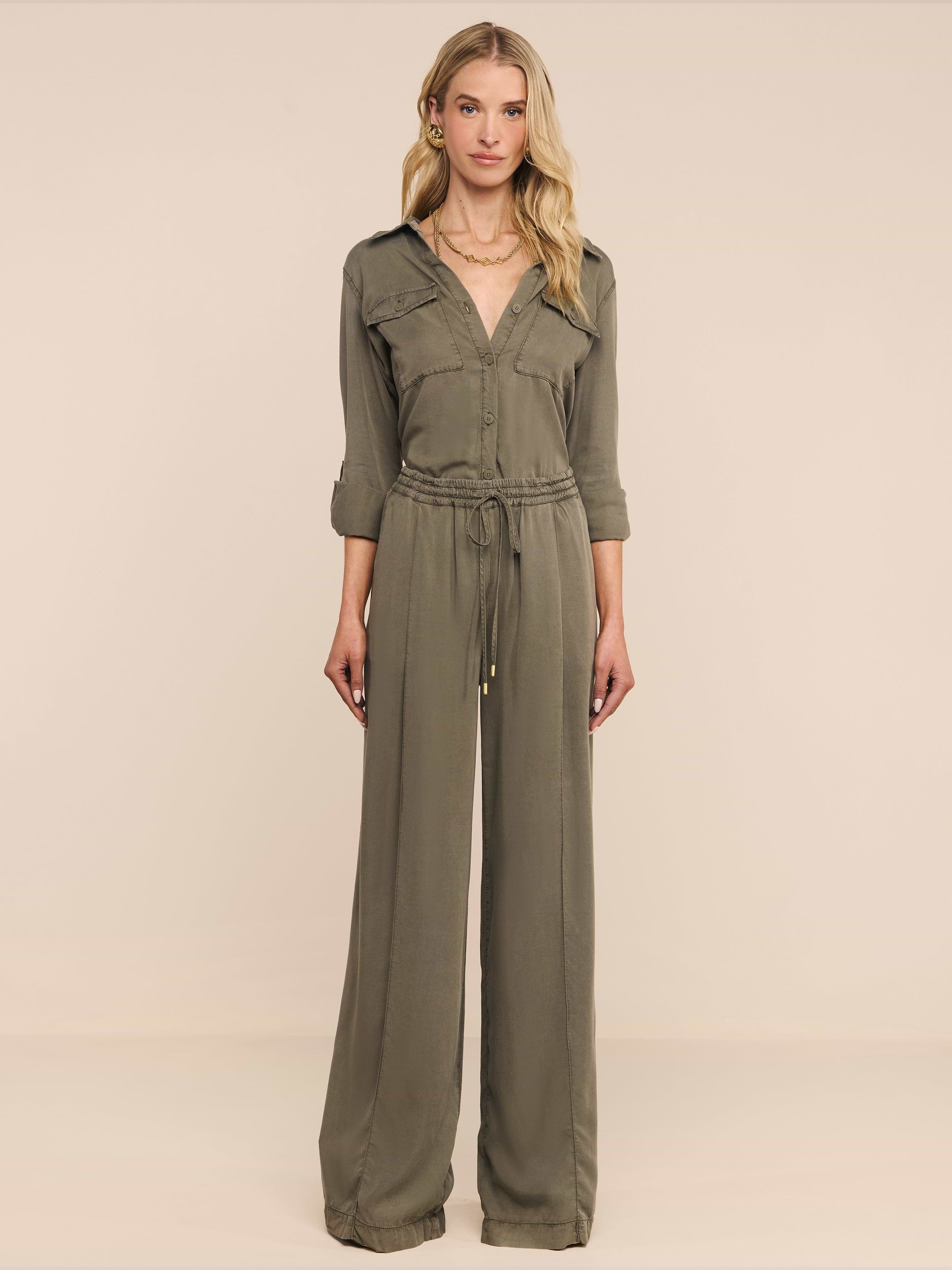 Una donna è rivolta in avanti e indossa i Connie Drawstring Trouser in verde oliva con coulisse in vita e pantaloni a gamba larga, su uno sfondo beige.