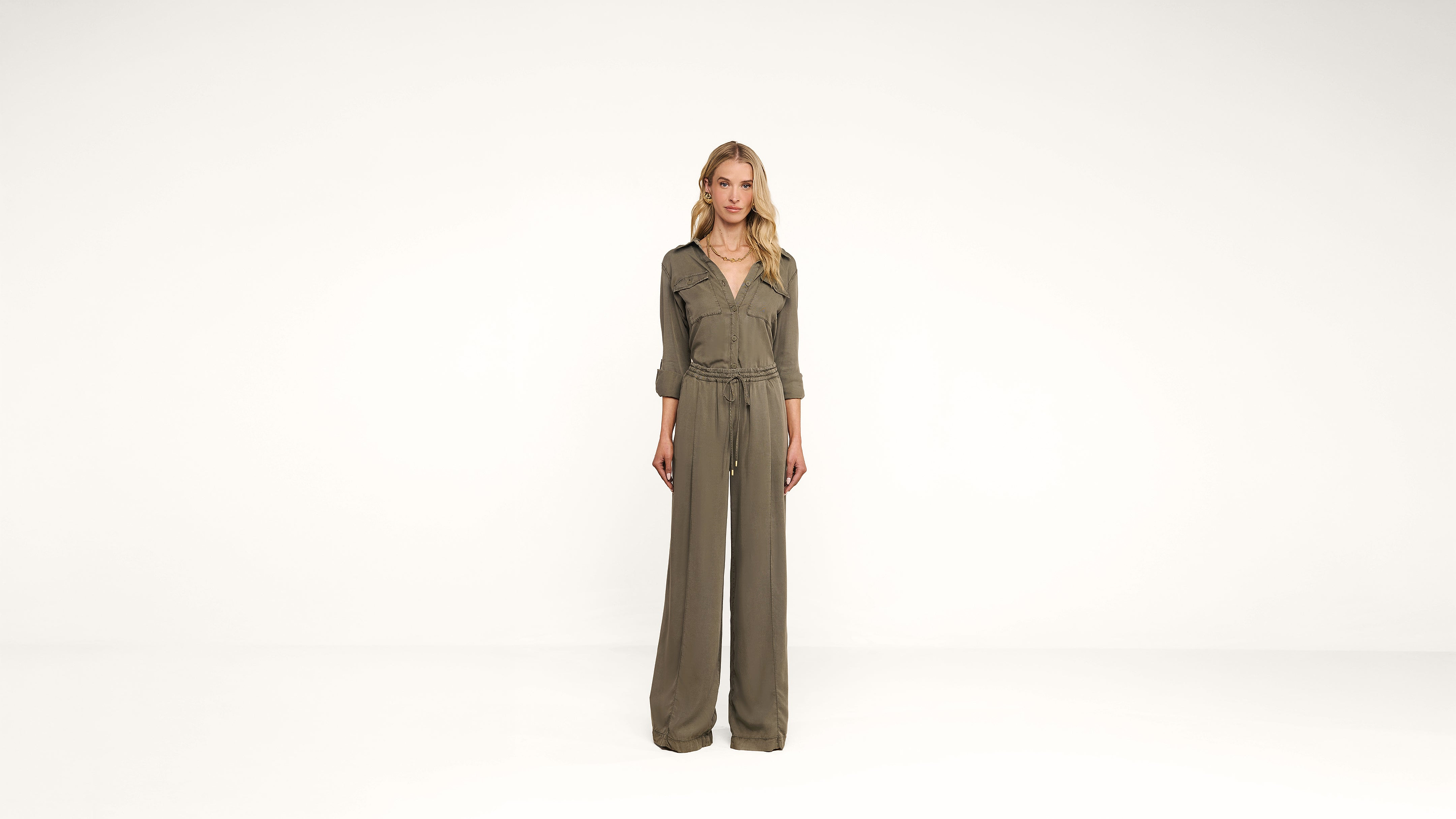 A woman stands against a plain white background wearing a khaki green jumpsuit with long, wide-leg pants and sleeves rolled to the elbows. She has blonde hair and a neutral expression.
