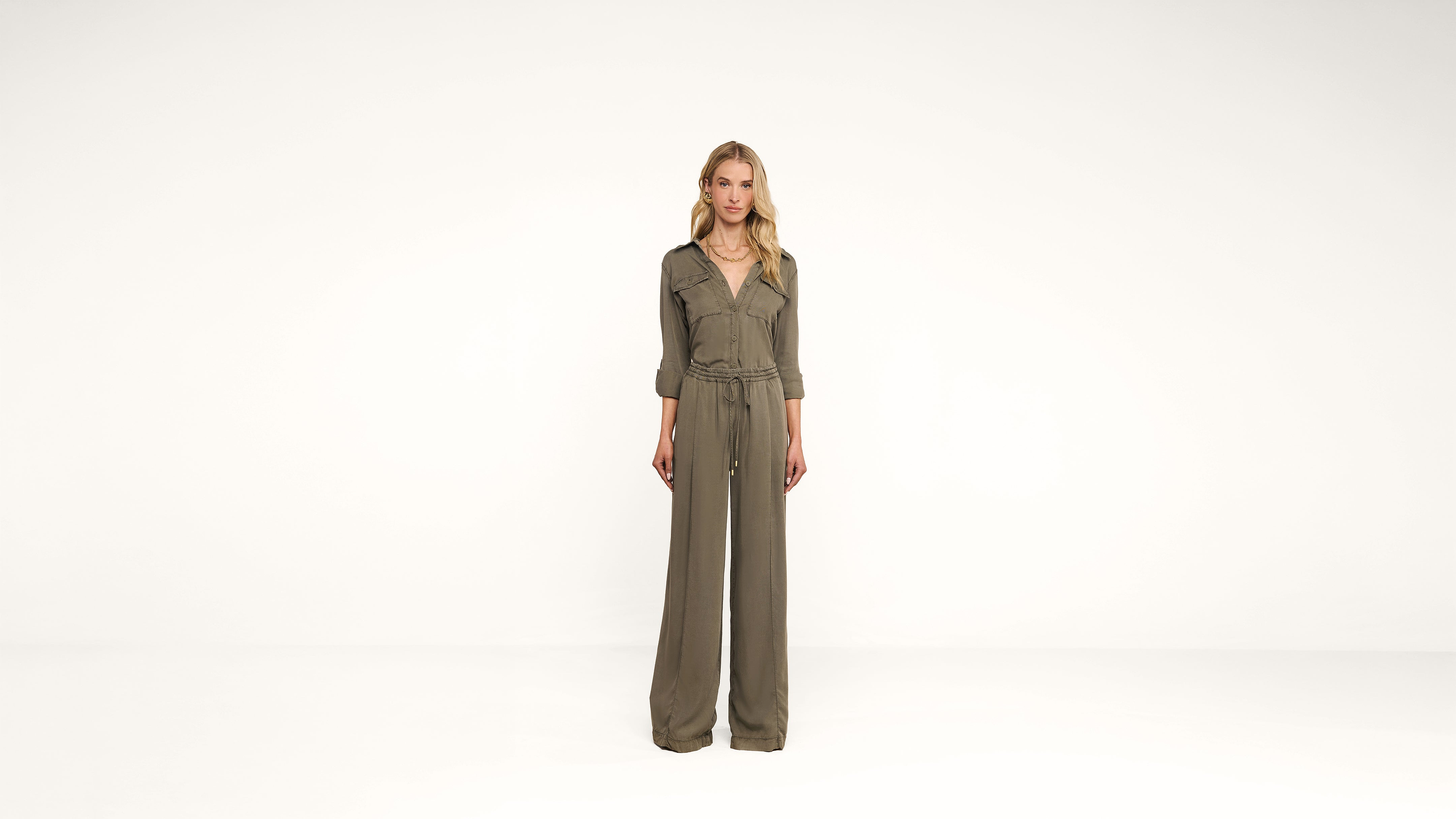 A woman stands against a plain white background wearing a khaki green jumpsuit with long, wide-leg pants and sleeves rolled to the elbows. She has blonde hair and a neutral expression.