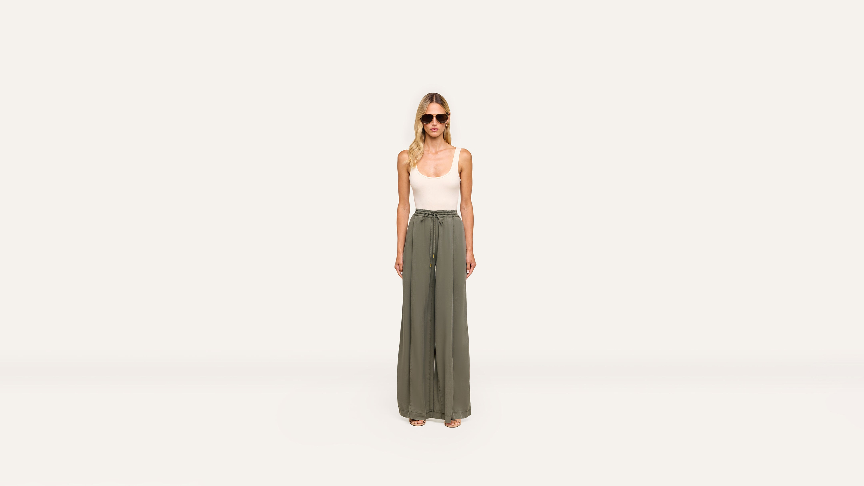 A woman stands against a plain white background, wearing sunglasses, a white sleeveless top, and high-waisted, wide-leg olive green pants for an effortlessly chic look.