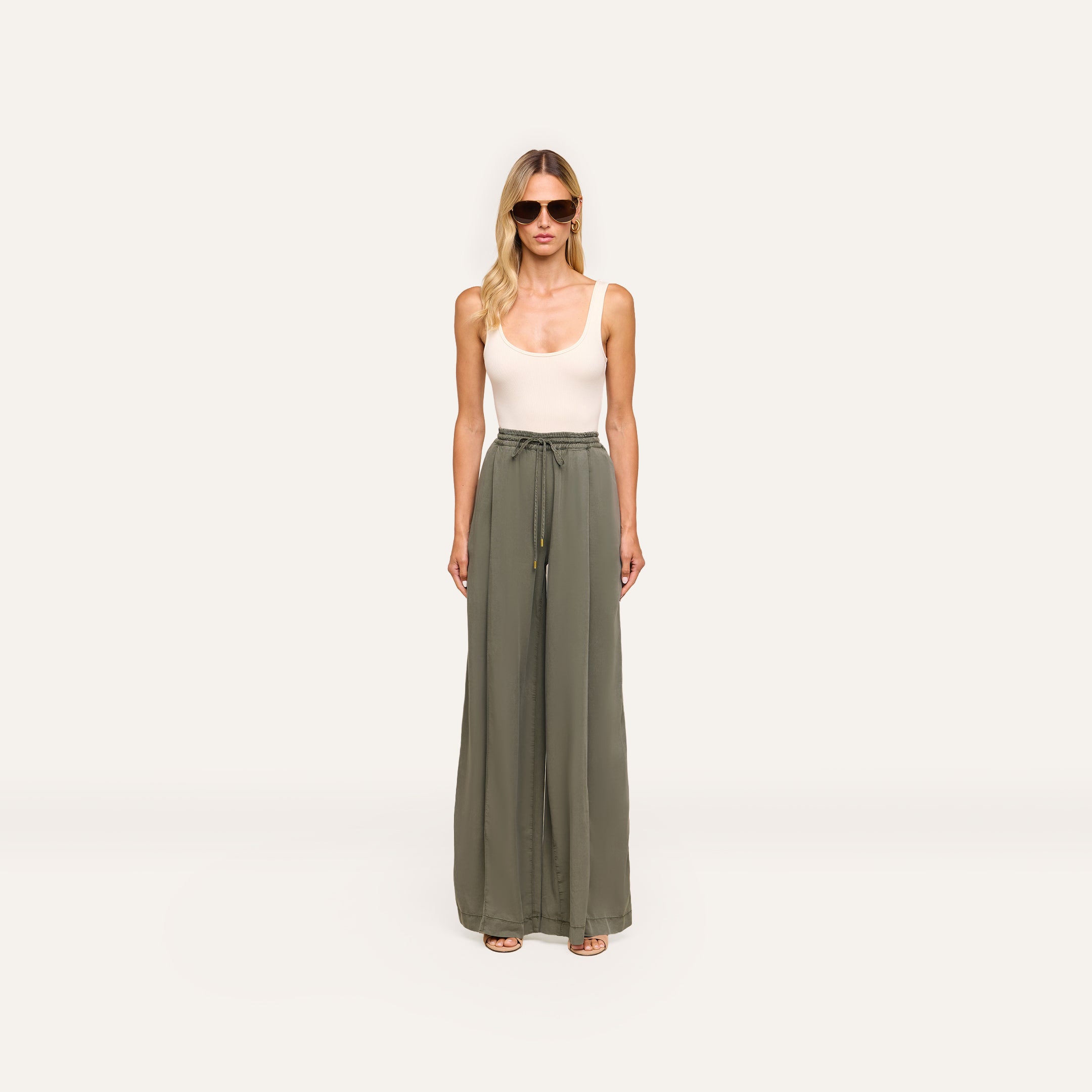 A woman in sunglasses and a white sleeveless top faces forward against a plain white background, wearing high-waisted olive green Connie Drawstring Trouser with a drawstring waistband.