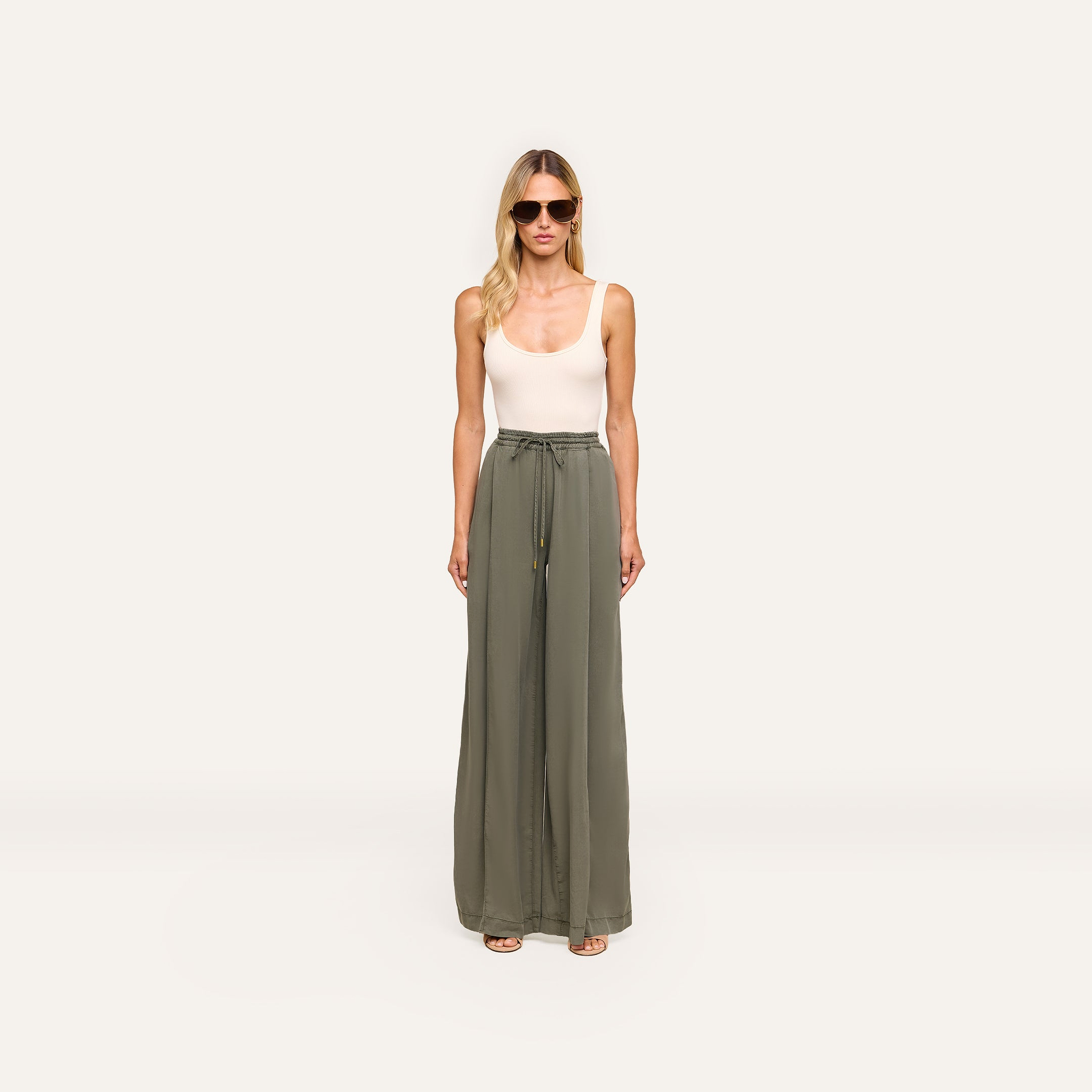 A woman in sunglasses and a white sleeveless top faces forward against a plain white background, wearing high-waisted olive green Connie Drawstring Trouser with a drawstring waistband.