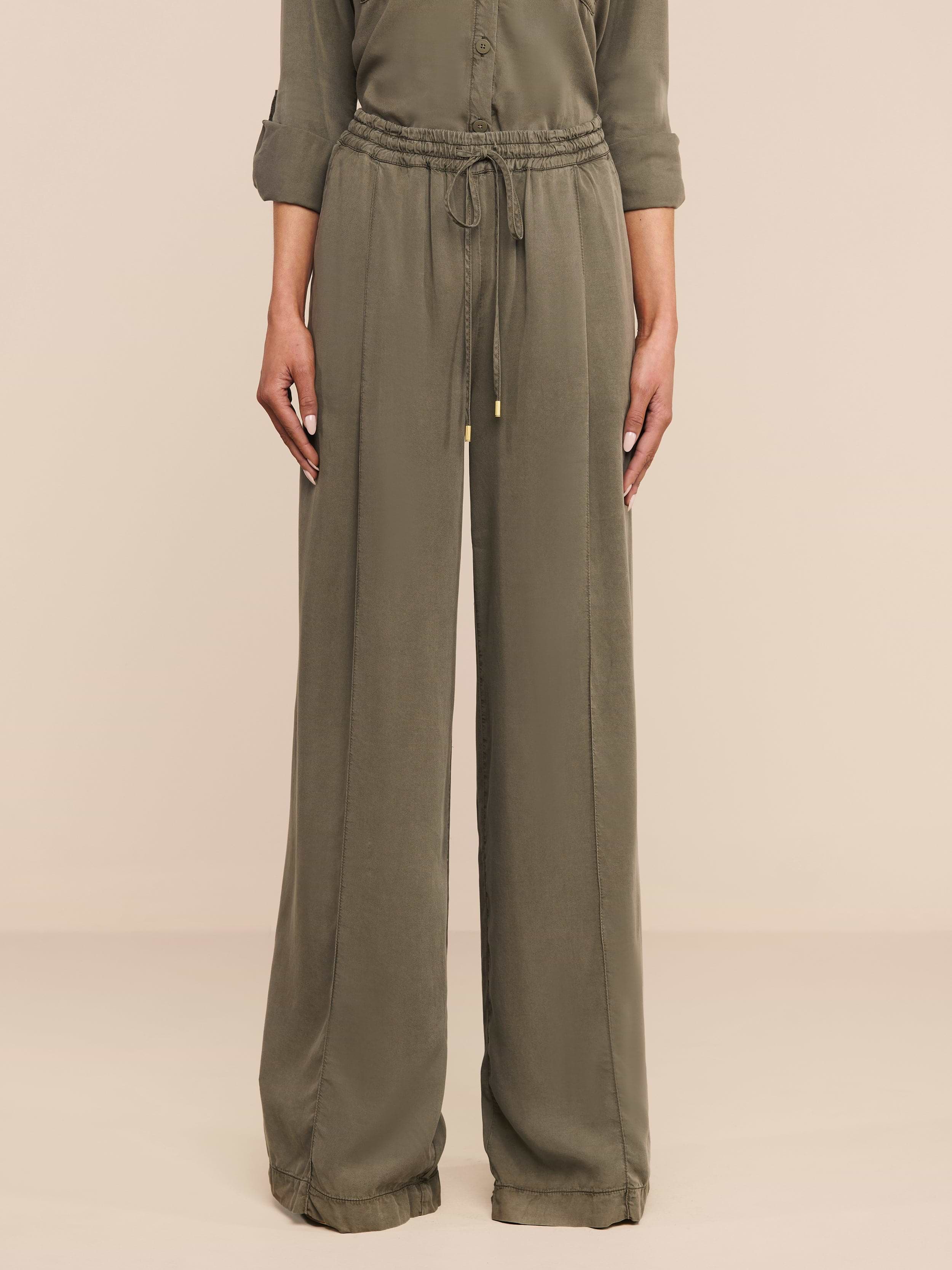 Indossa i Connie Drawstring Trouser in twill verde oliva con vestibilità rilassata, coulisse in vita e polsini arrotolati; la modella è in piedi su uno sfondo neutro con la parte superiore del busto parzialmente visibile.