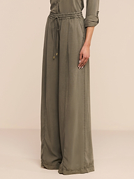 Una persona che indossa i pantaloni Connie Drawstring Trouser in verde oliva con una camicia a maniche lunghe abbinata si trova su uno sfondo beige semplice.