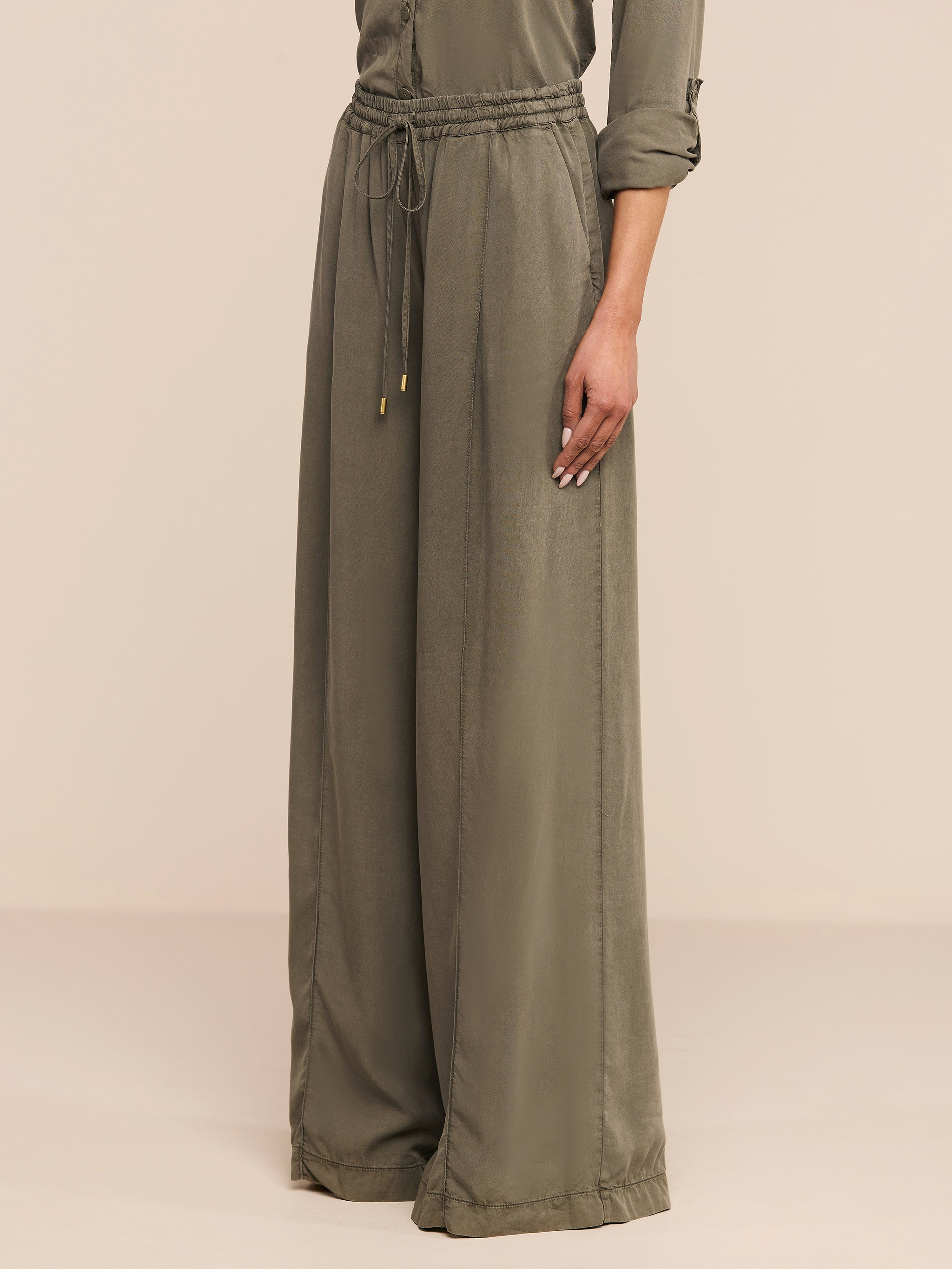 Una persona che indossa i pantaloni Connie Drawstring Trouser in verde oliva con una camicia a maniche lunghe abbinata si trova su uno sfondo beige semplice.