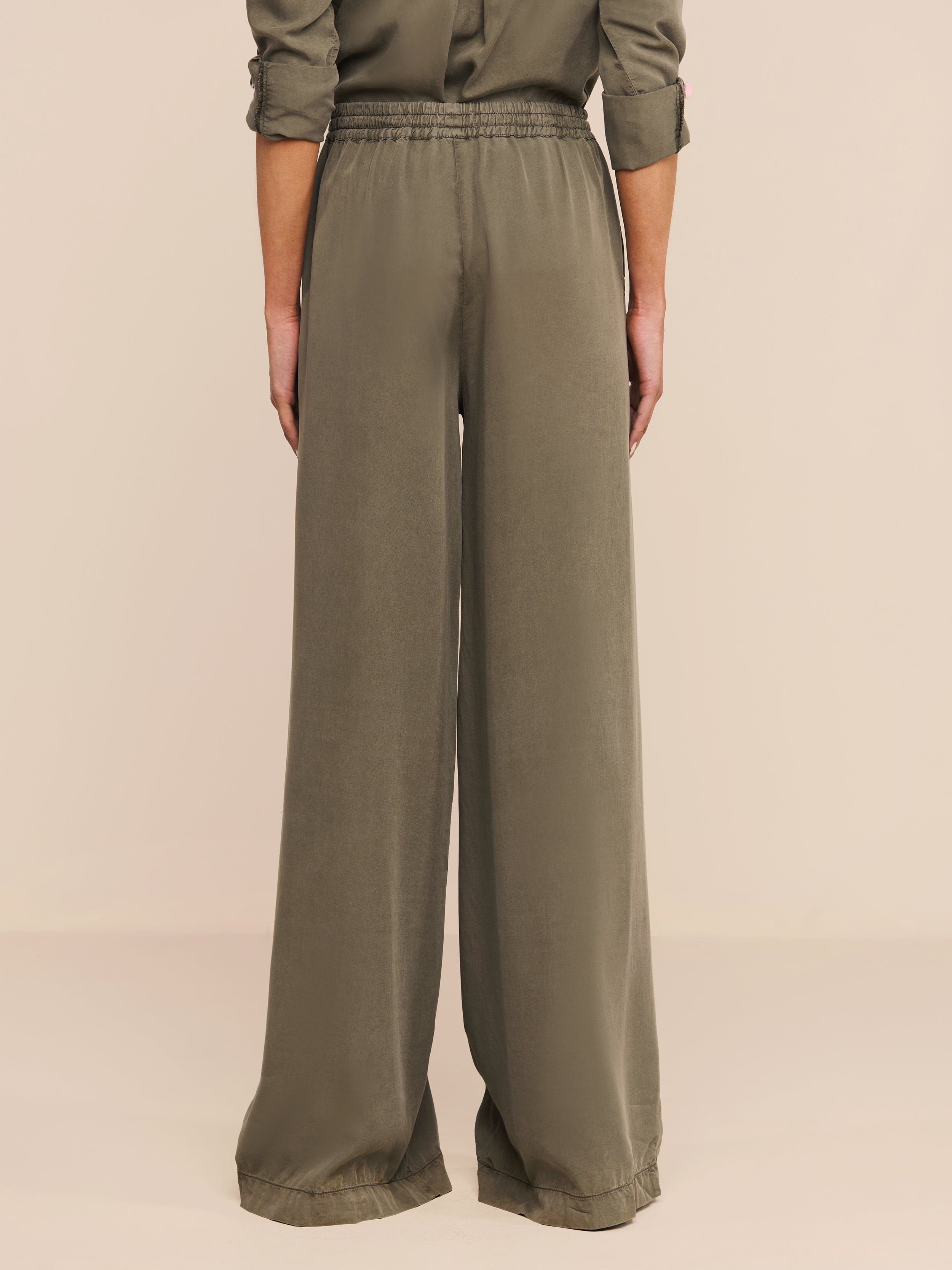 La persona si trova di spalle alla telecamera e indossa pantaloni larghi Connie Drawstring Trouser in twill verde oliva e una camicia a maniche lunghe abbinata con maniche arrotolate. Lo sfondo è semplice e neutro.