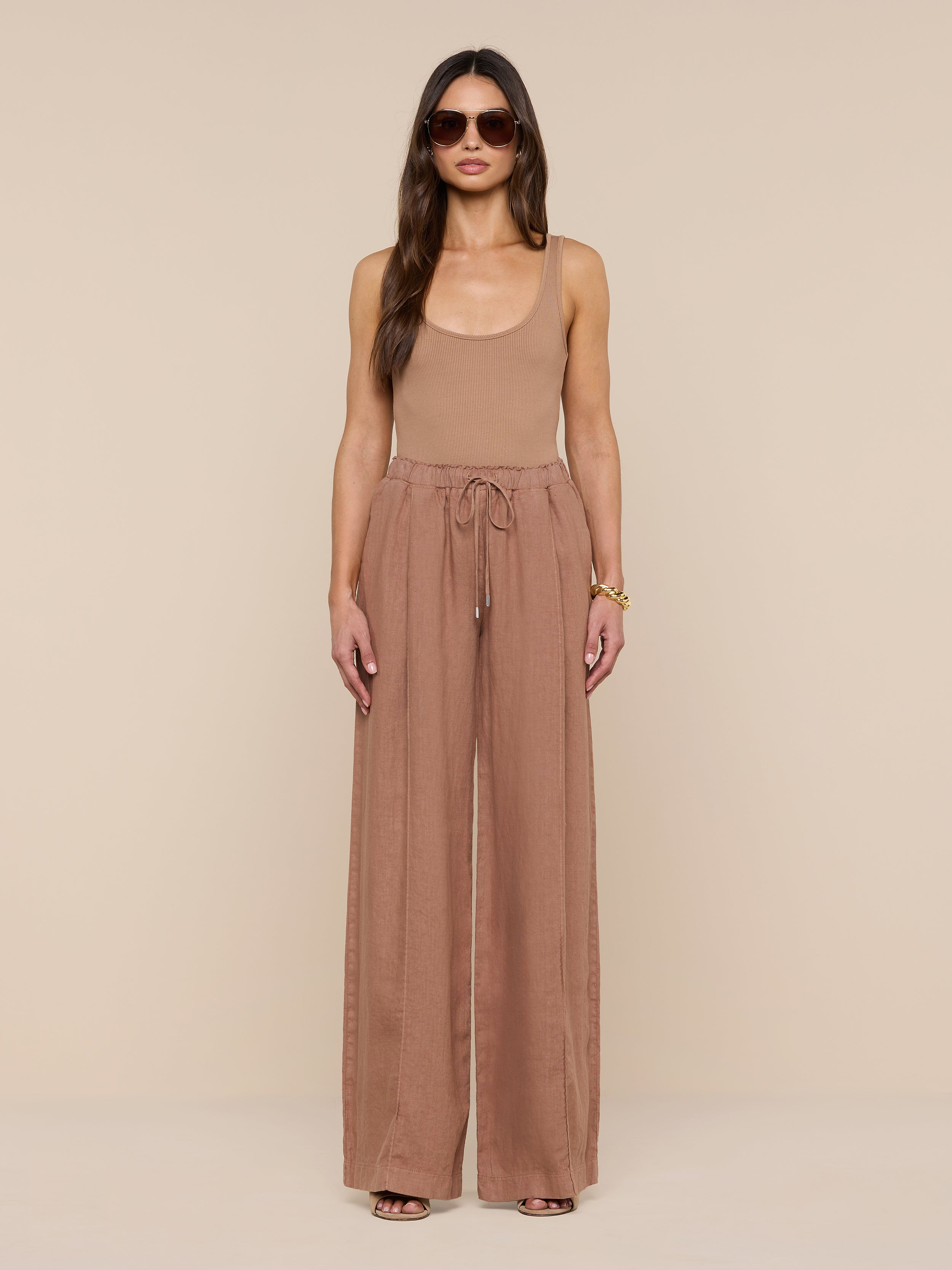 A woman stands facing forward against a plain white background, wearing sunglasses, a tan sleeveless top, and the Connie Linen Drawstring Trouser—loose, high-waisted linen pants with a drawstring waistband.
