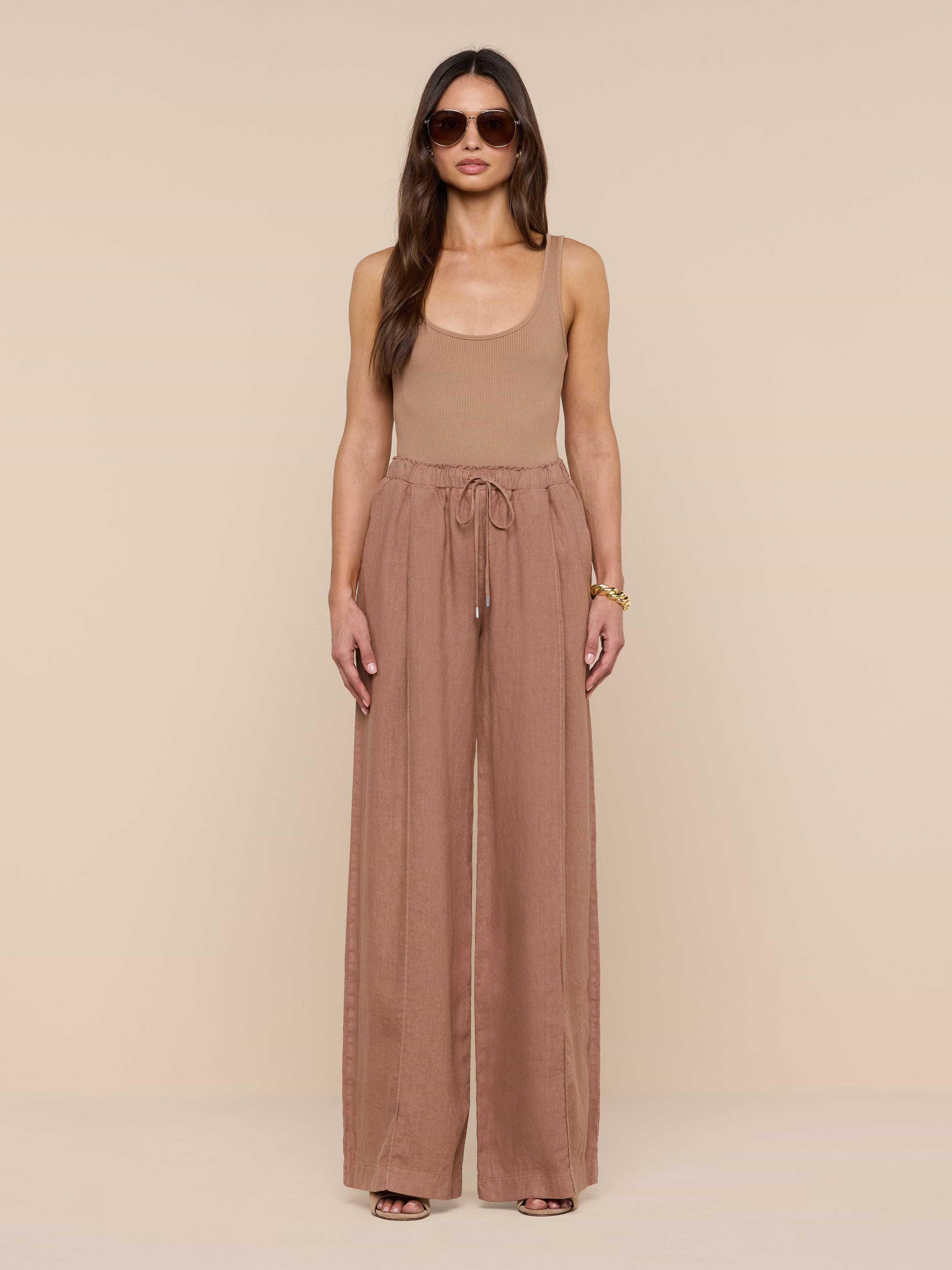 A woman stands facing forward against a plain white background, wearing sunglasses, a tan sleeveless top, and the Connie Linen Drawstring Trouser—loose, high-waisted linen pants with a drawstring waistband.