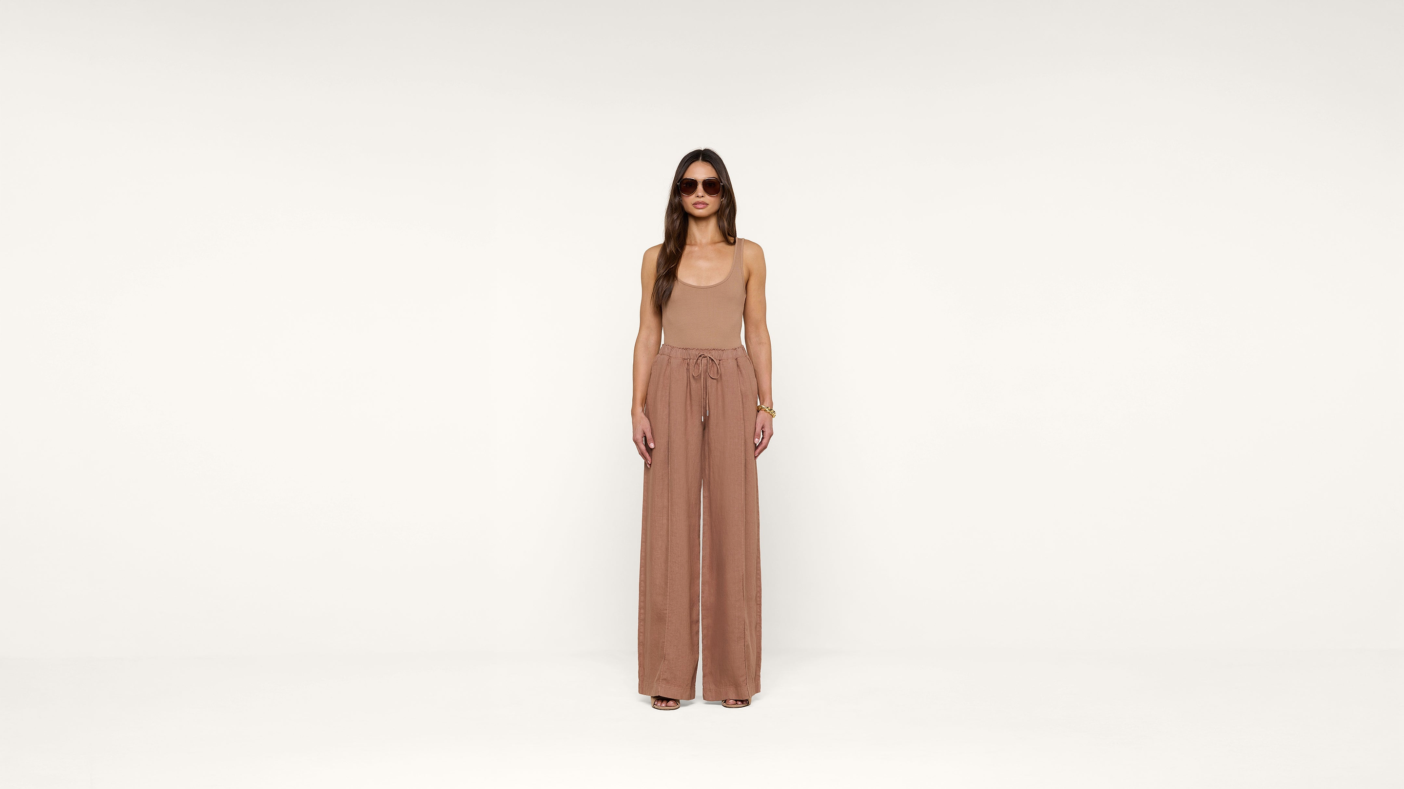 A woman in sunglasses wearing a sleeveless tan jumpsuit with wide legs stands against a plain white background, looking directly at the camera.
