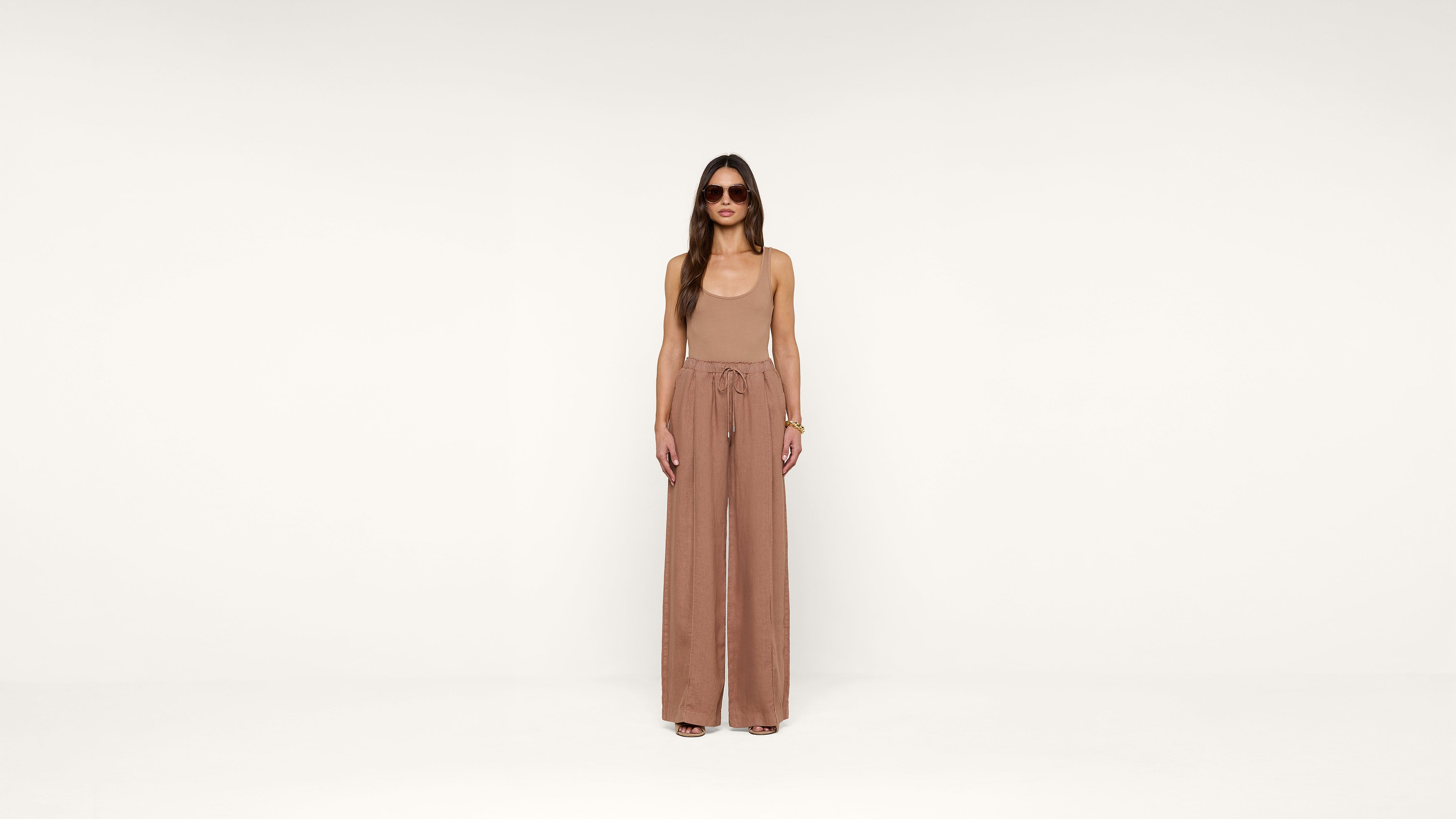 A woman in sunglasses wearing a sleeveless tan jumpsuit with wide legs stands against a plain white background, looking directly at the camera.