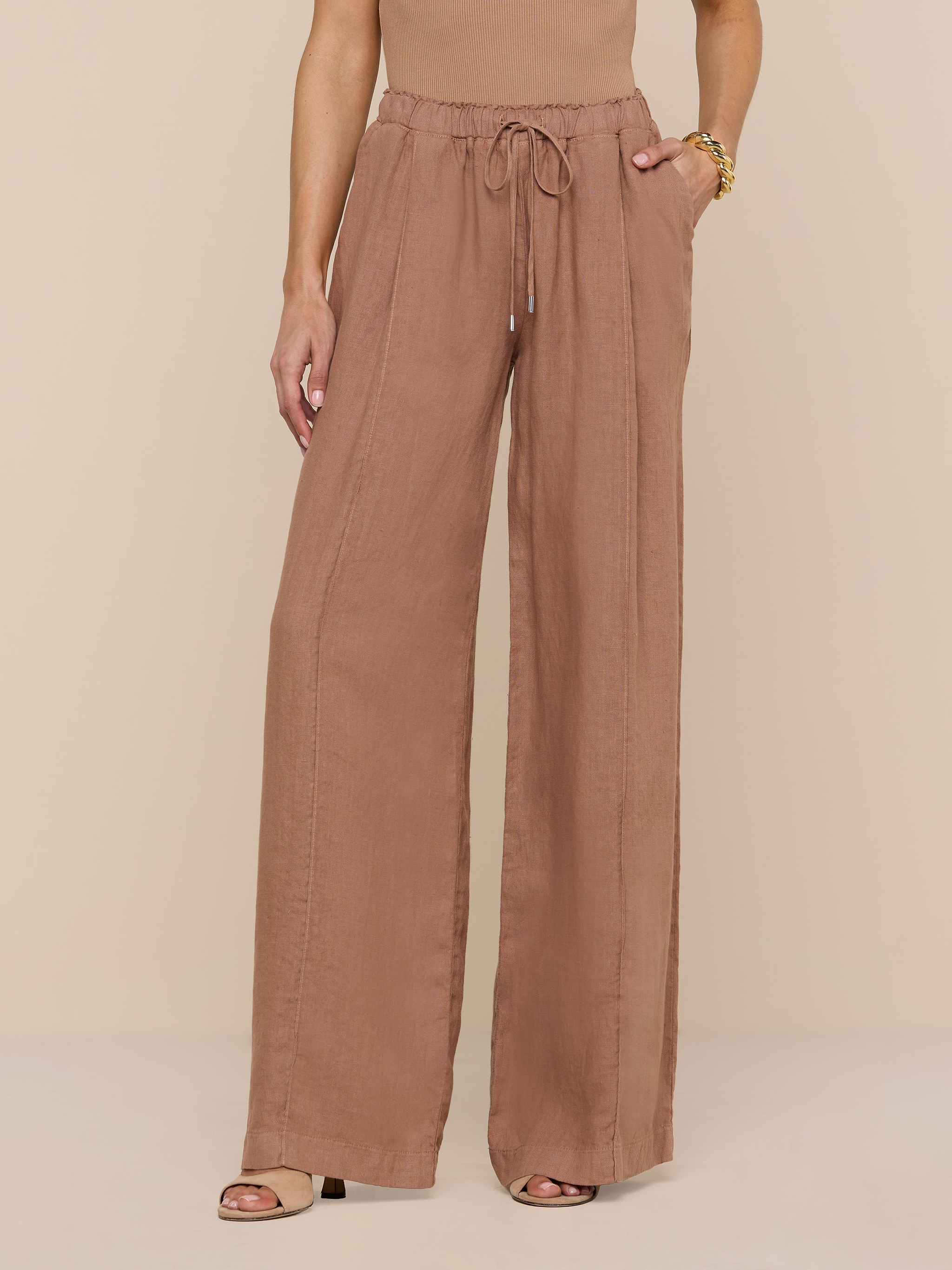 A person wearing the Connie Linen Drawstring Trouser—a wide-leg, high-waisted, light brown linen pant with a drawstring waistband—paired with a sleeveless top and heeled sandals, stands against a plain white background, with only the lower half of their body visible.