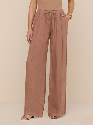 Model wears the Connie Linen Drawstring Trouser—high-waisted, wide-leg linen pants with a drawstring waistband and side pockets—paired with open-toe heels and a sleeveless top on a beige background.