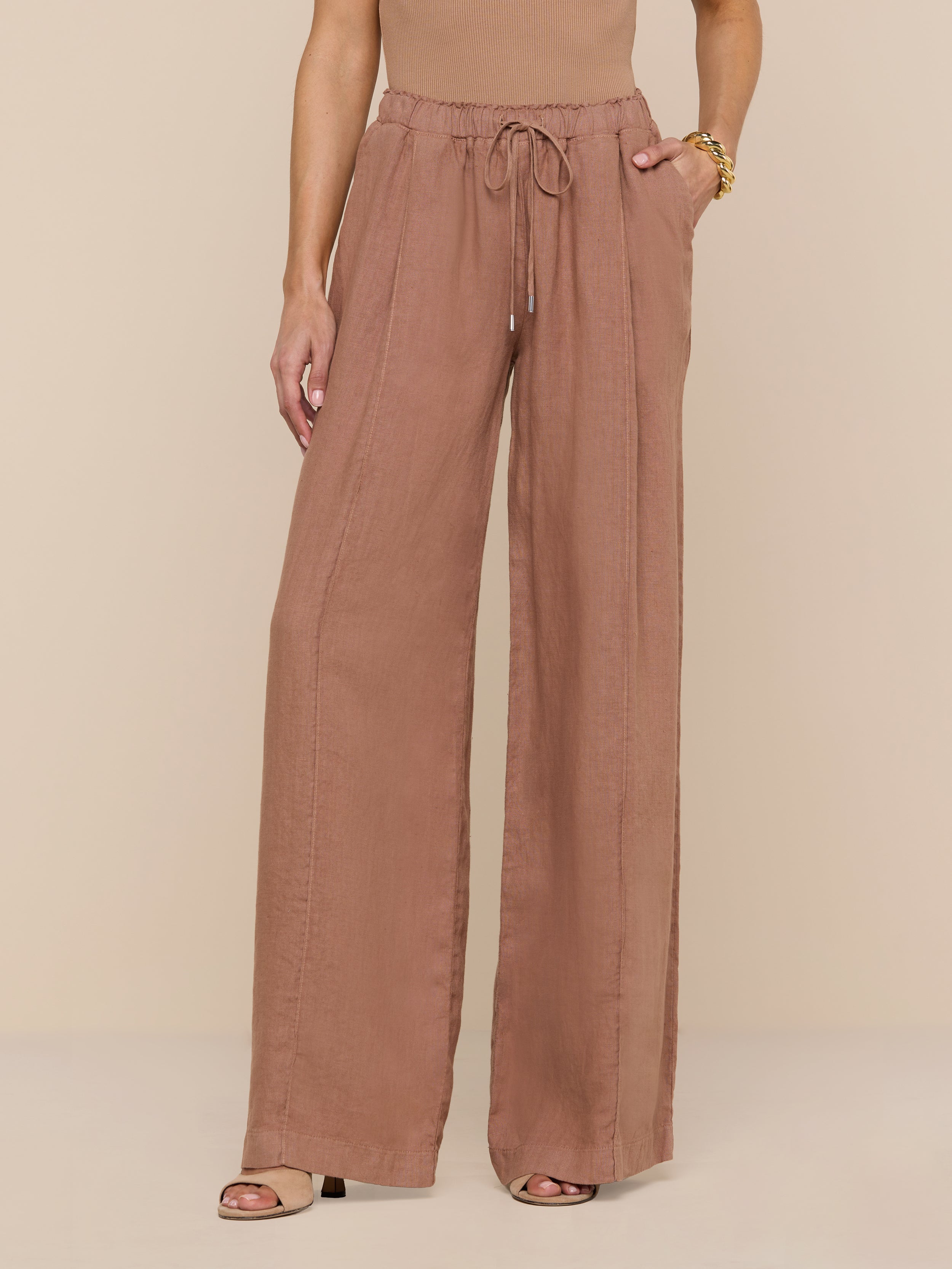 A person wearing the Connie Linen Drawstring Trouser—a wide-leg, high-waisted, light brown linen pant with a drawstring waistband—paired with a sleeveless top and heeled sandals, stands against a plain white background, with only the lower half of their body visible.