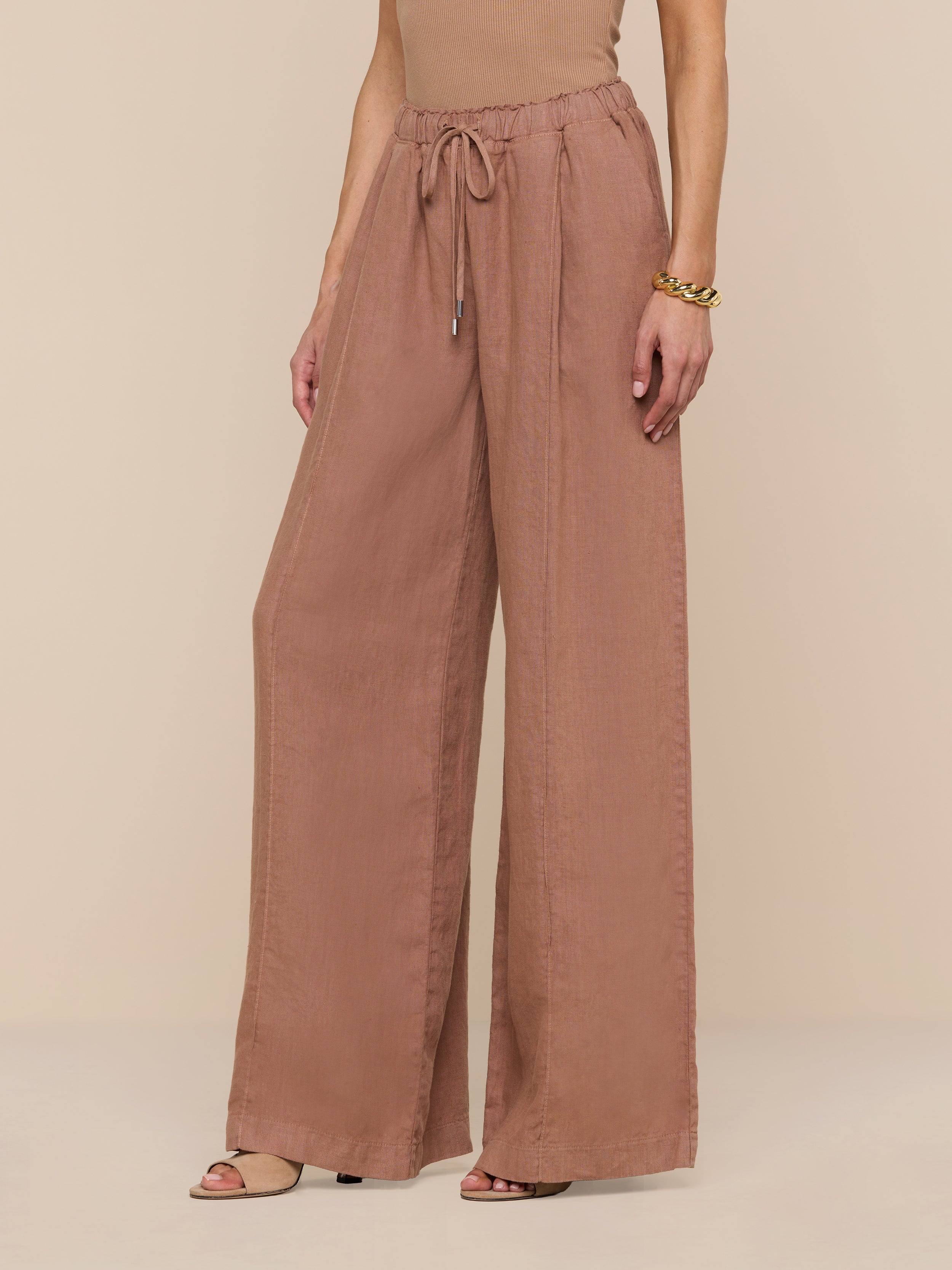 A person wearing the Connie Linen Drawstring Trouser—loose, wide-leg, brown linen pants featuring a drawstring waistband—paired with a sleeveless top and sandals, stands against a plain white background.