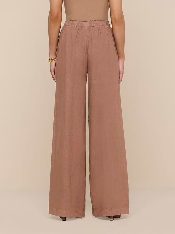 Standing against a white background, the person wears high-waisted, wide-leg Connie Linen Drawstring Trouser in brown paired with a sleeveless top. The view from the back highlights the full length of these vacation-ready pants.