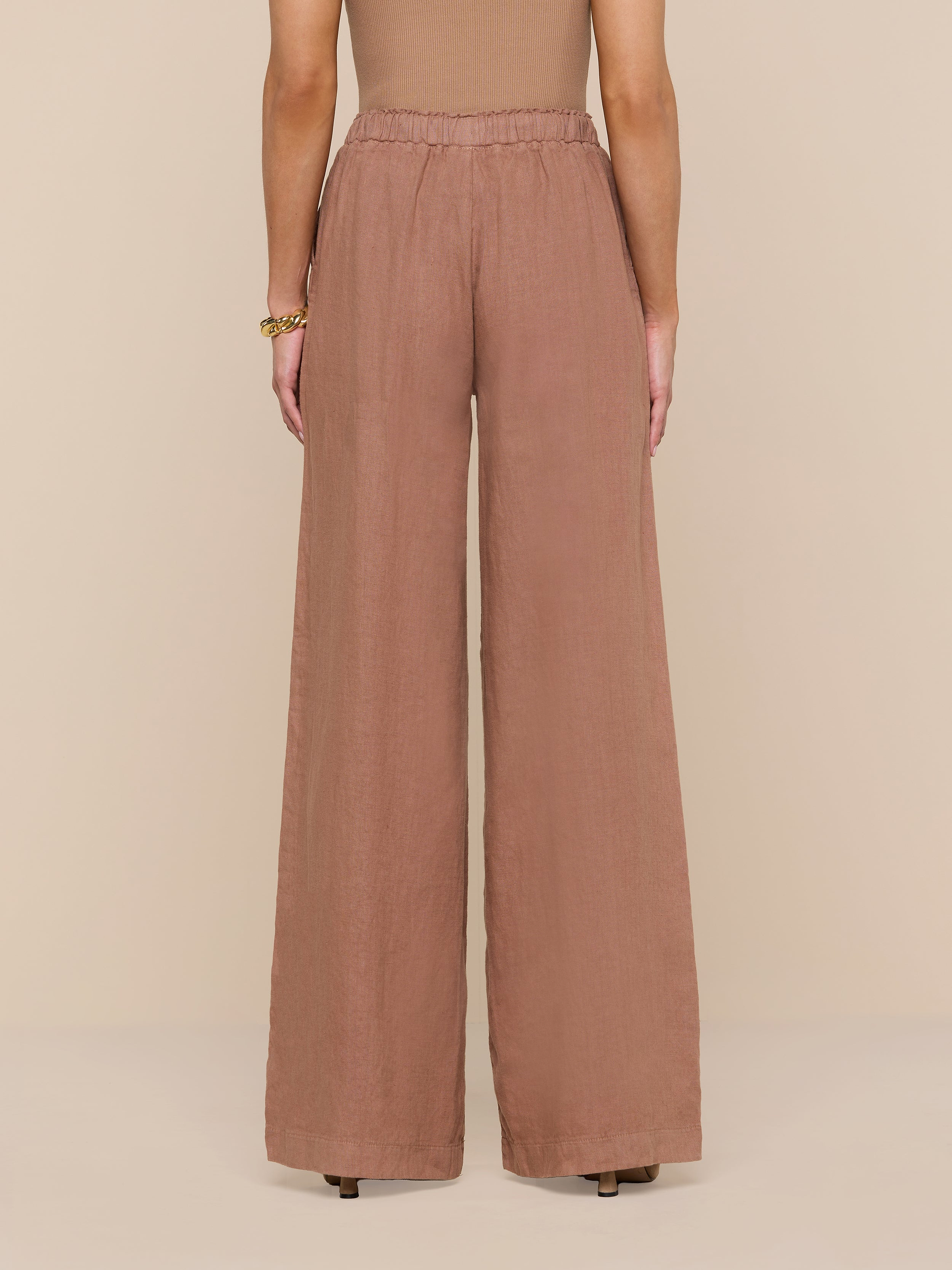 Standing against a white background, the person wears high-waisted, wide-leg Connie Linen Drawstring Trouser in brown paired with a sleeveless top. The view from the back highlights the full length of these vacation-ready pants.