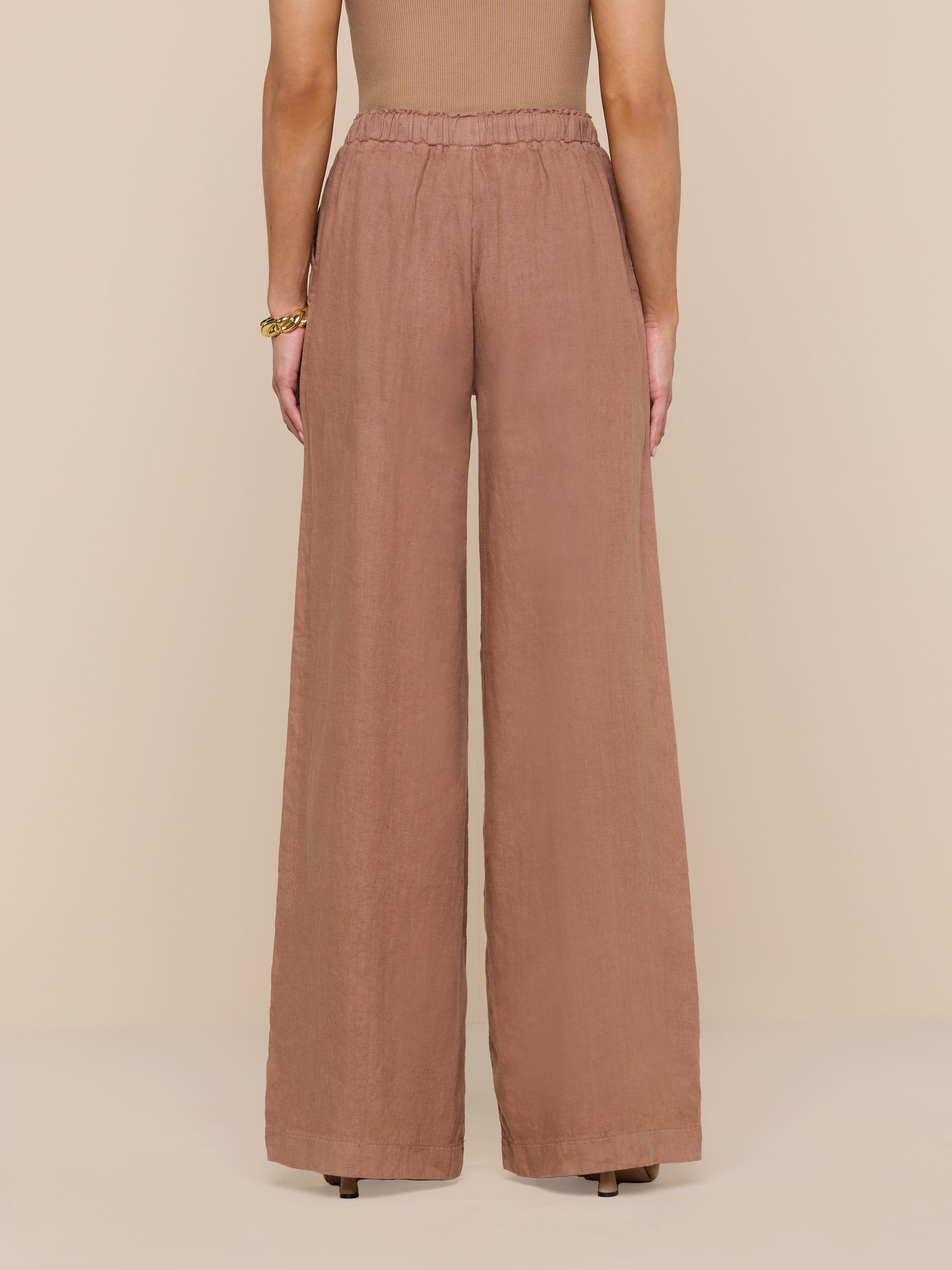 Standing against a white background, the person wears high-waisted, wide-leg Connie Linen Drawstring Trouser in brown paired with a sleeveless top. The view from the back highlights the full length of these vacation-ready pants.