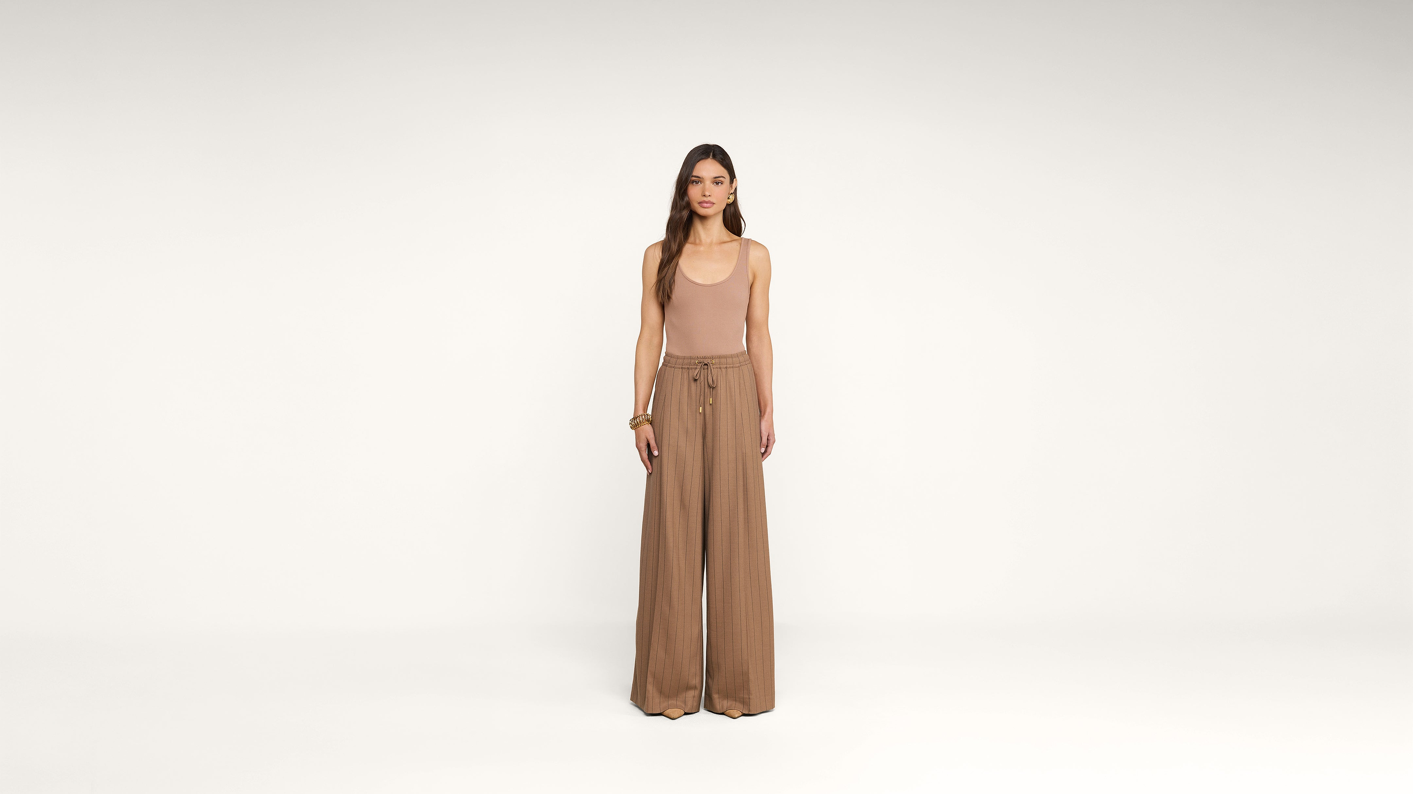 A woman stands facing forward, wearing a beige sleeveless top and high-waisted, wide-leg brown pants in a minimalist, white studio setting.