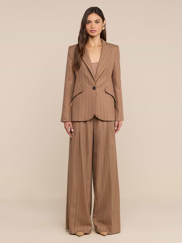 Une femme aux longs cheveux ondulés et au maquillage discret se tient debout, face à lobjectif, vêtue dun tailleur à fines rayures beige clair composé dun pantalon « Connie Drawstring » et dun blazer à un bouton, posant devant un fond beige uni.