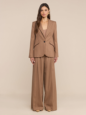 A woman with long, wavy hair and neutral makeup stands facing forward in a tan pinstripe suit featuring the Connie Drawstring Trouser and a single-button blazer, posed against a plain beige background.